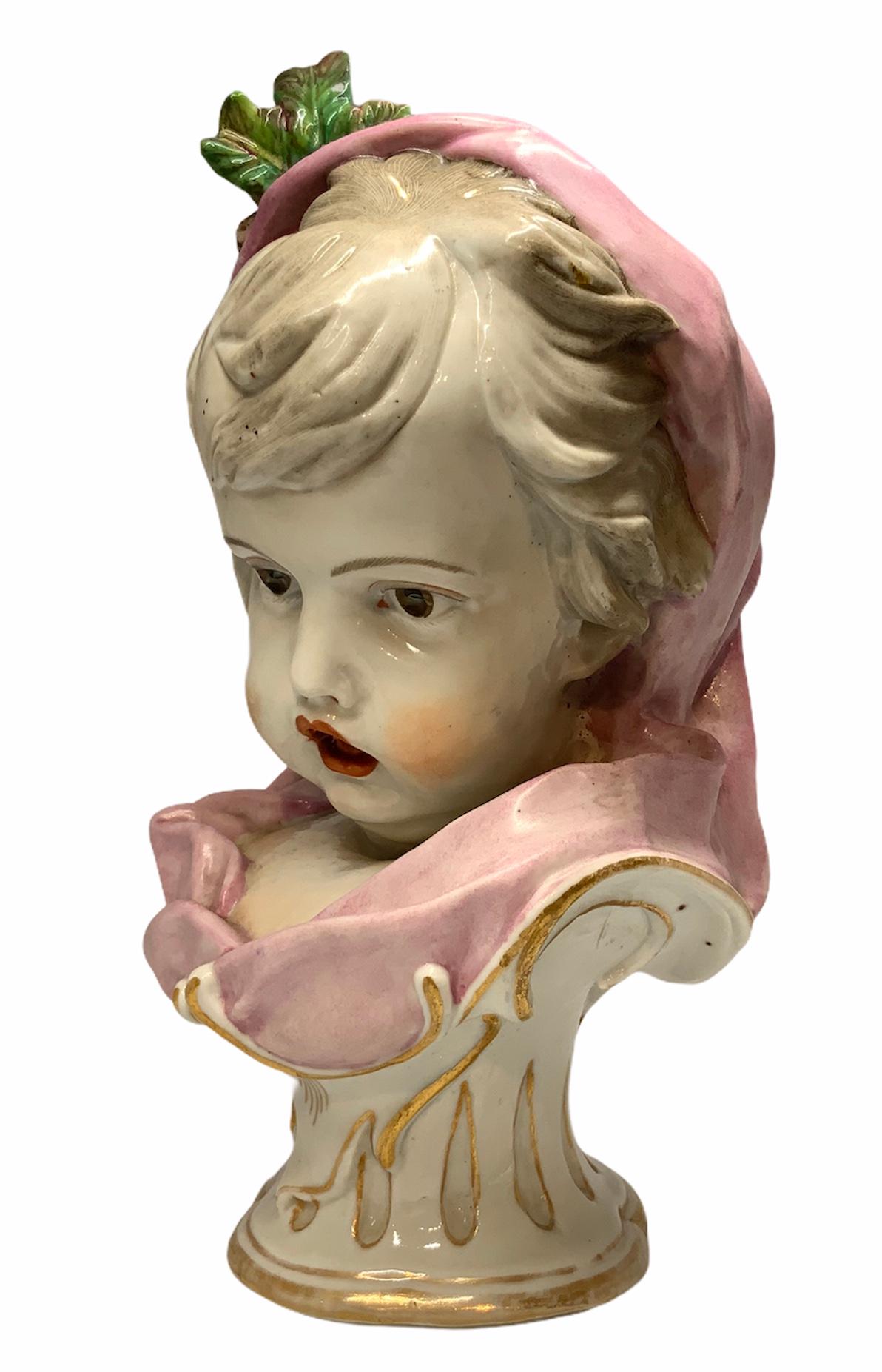 Depicting a bust of a baby girl whose ash hair head and upper chest is covered by a pink waved cape. This is adorned with pine nuts and small pine branches in the top of the head. She has deep brown eyes, red lips, and prominent cheek bones. It