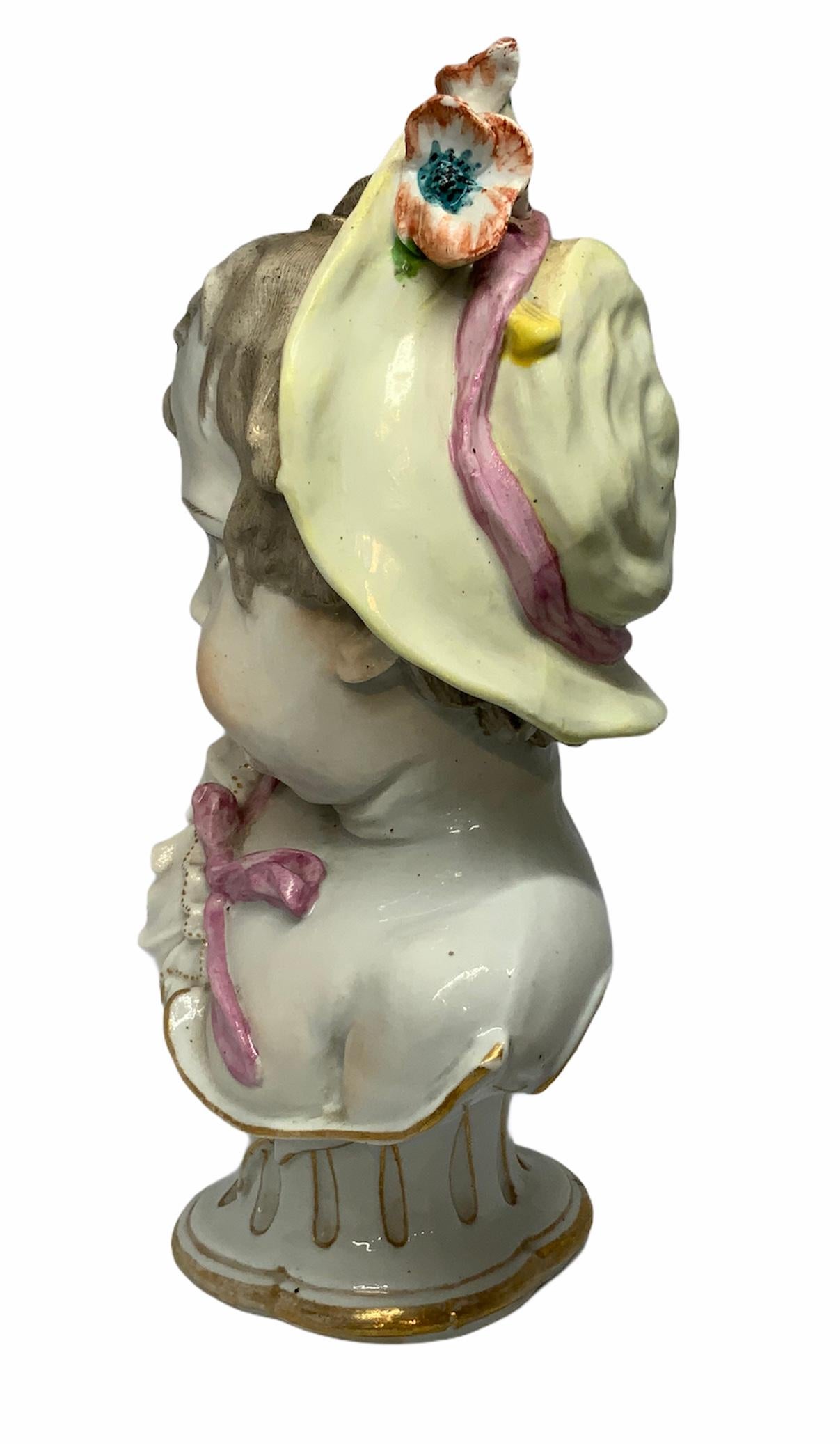 Depicting a bust of a baby girl with an innocent gaze, red lips, and uncombed blond hair. She is wearing a gilt white ruffle top and in her head a charming pink bow hat with flowers. It stands on a gilt and white quatrefoile base. There are two blue