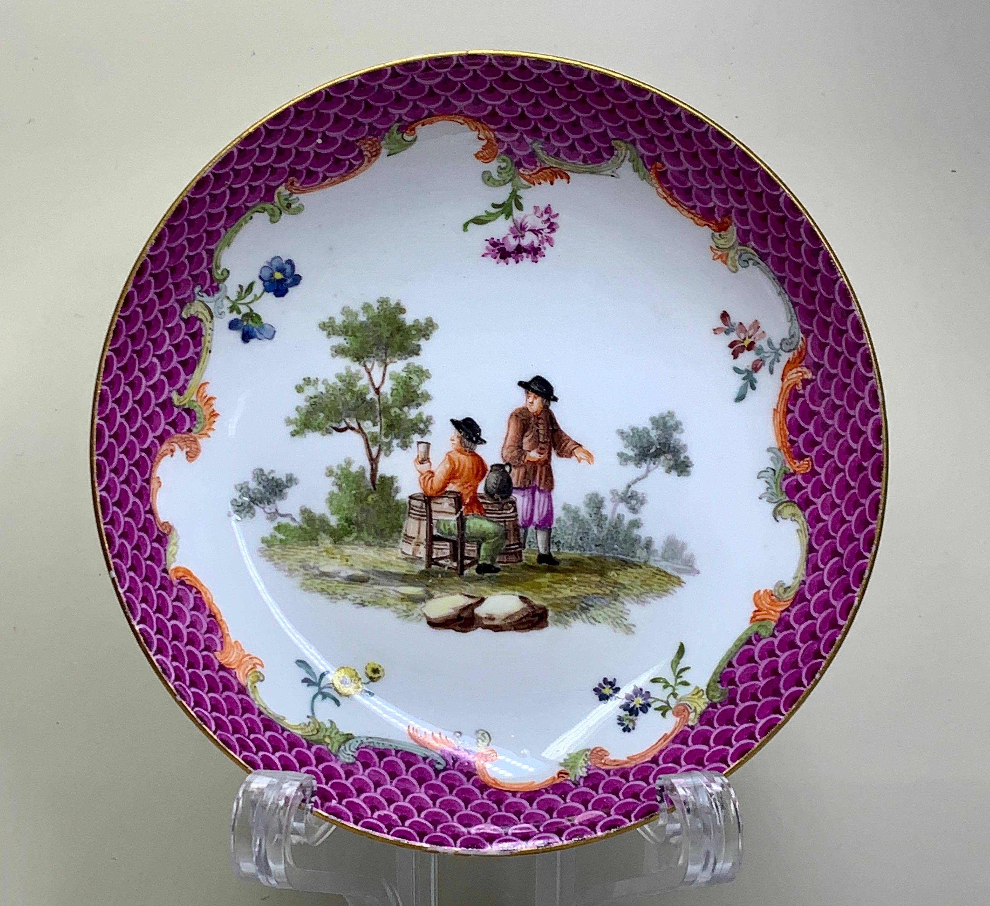 A quality Meissen teabowl and saucer, superbly painted with Watteauesque scenes in a garden, superb pink or purple scale trim all around the rims with nice scroll work underneath. There is some gilding to the edges., both pieces have under glazed