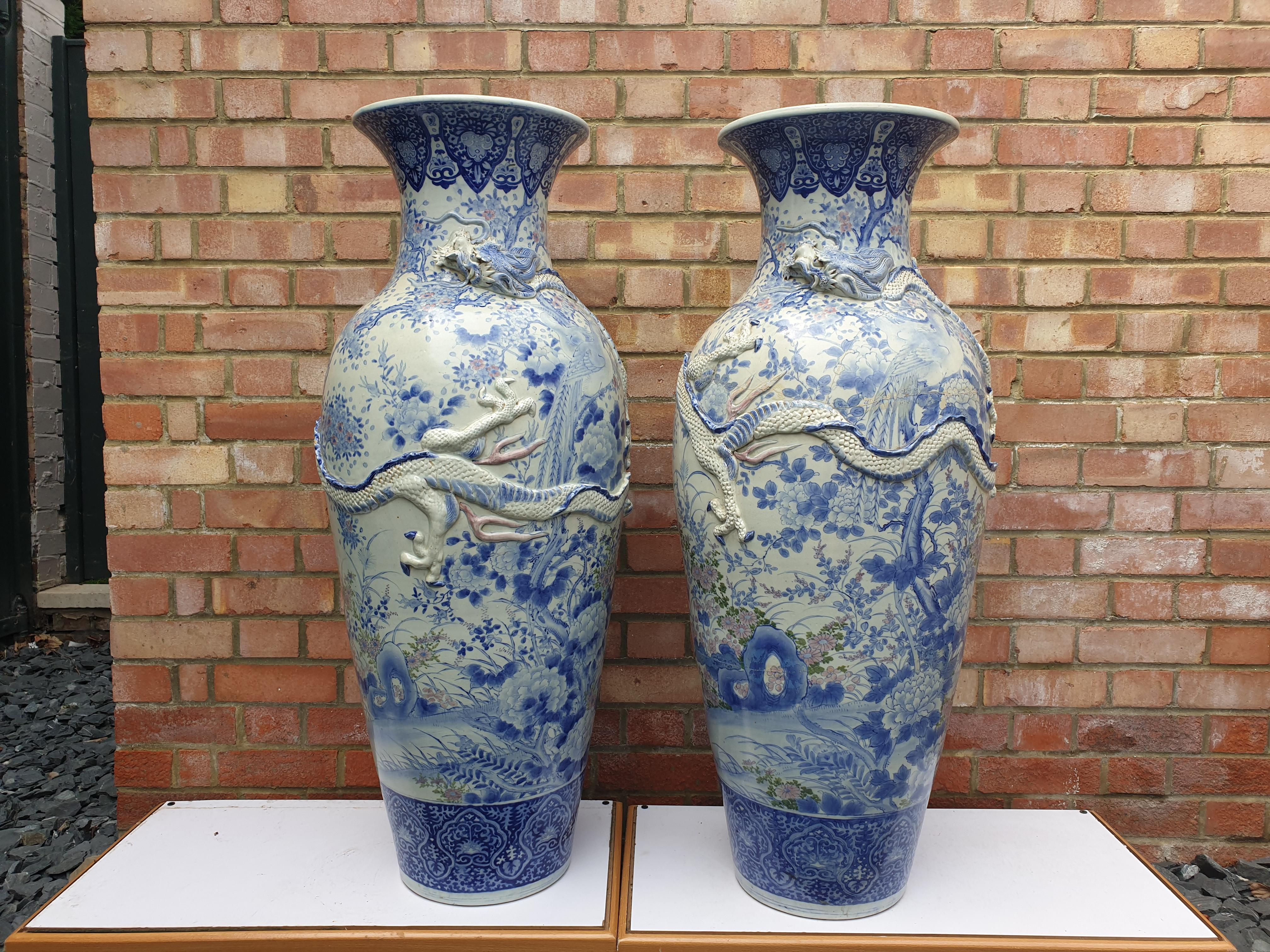 An impressive pair of Japanese vases standing upright with a short neck and a turned out top rim or lip. In traditional colors of blue and white with embossed three claw molded dragons wrapped around the center body of the vases. Some flowers are