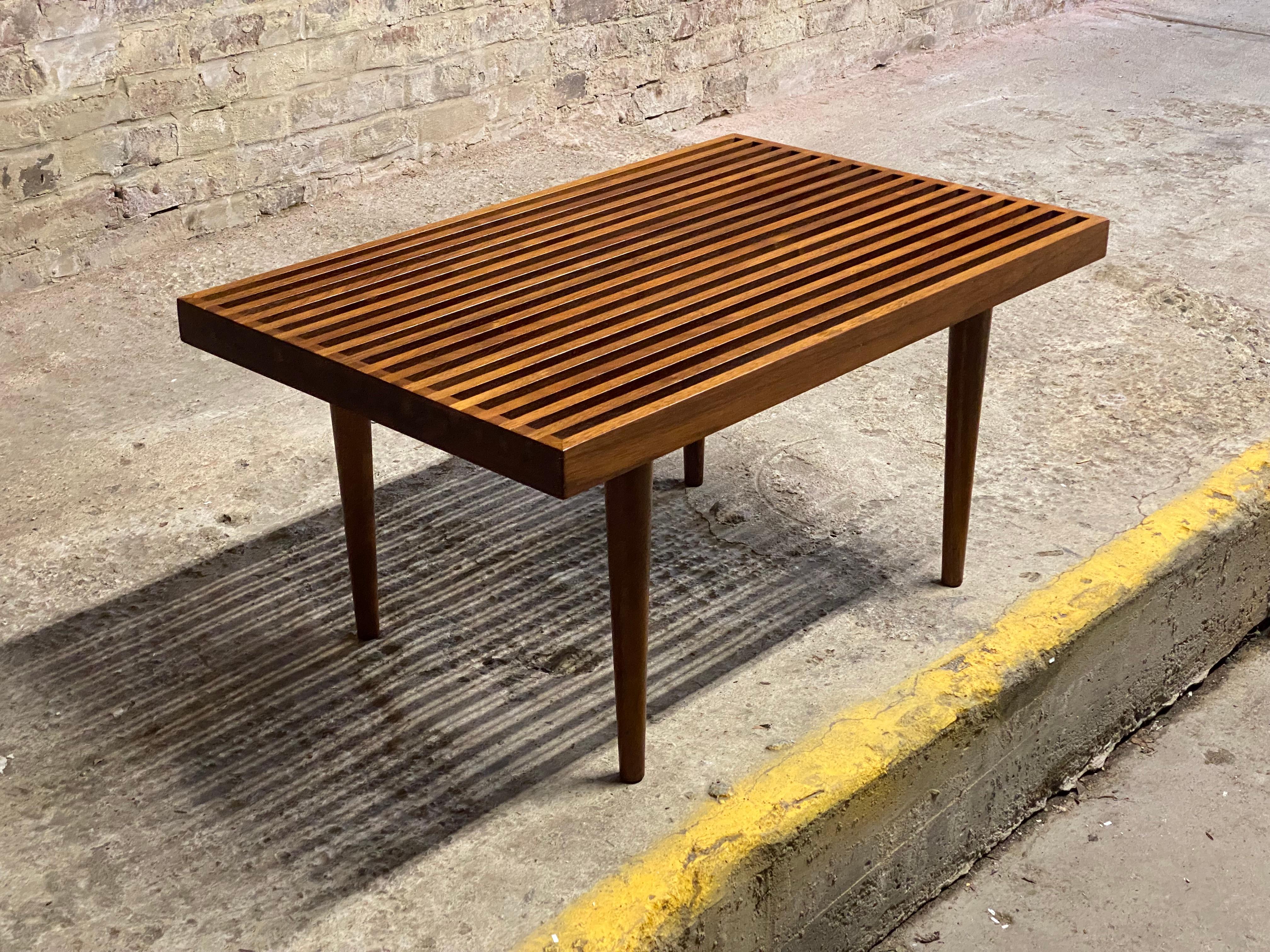 A Mel Smilow (1922-2002) designed solid walnut slat table. Wonderful oiled walnut featuring that signature splined corners, cross cut color contrast sides and removable tapered legs. Flat pack design. These are great accent tables or small benches