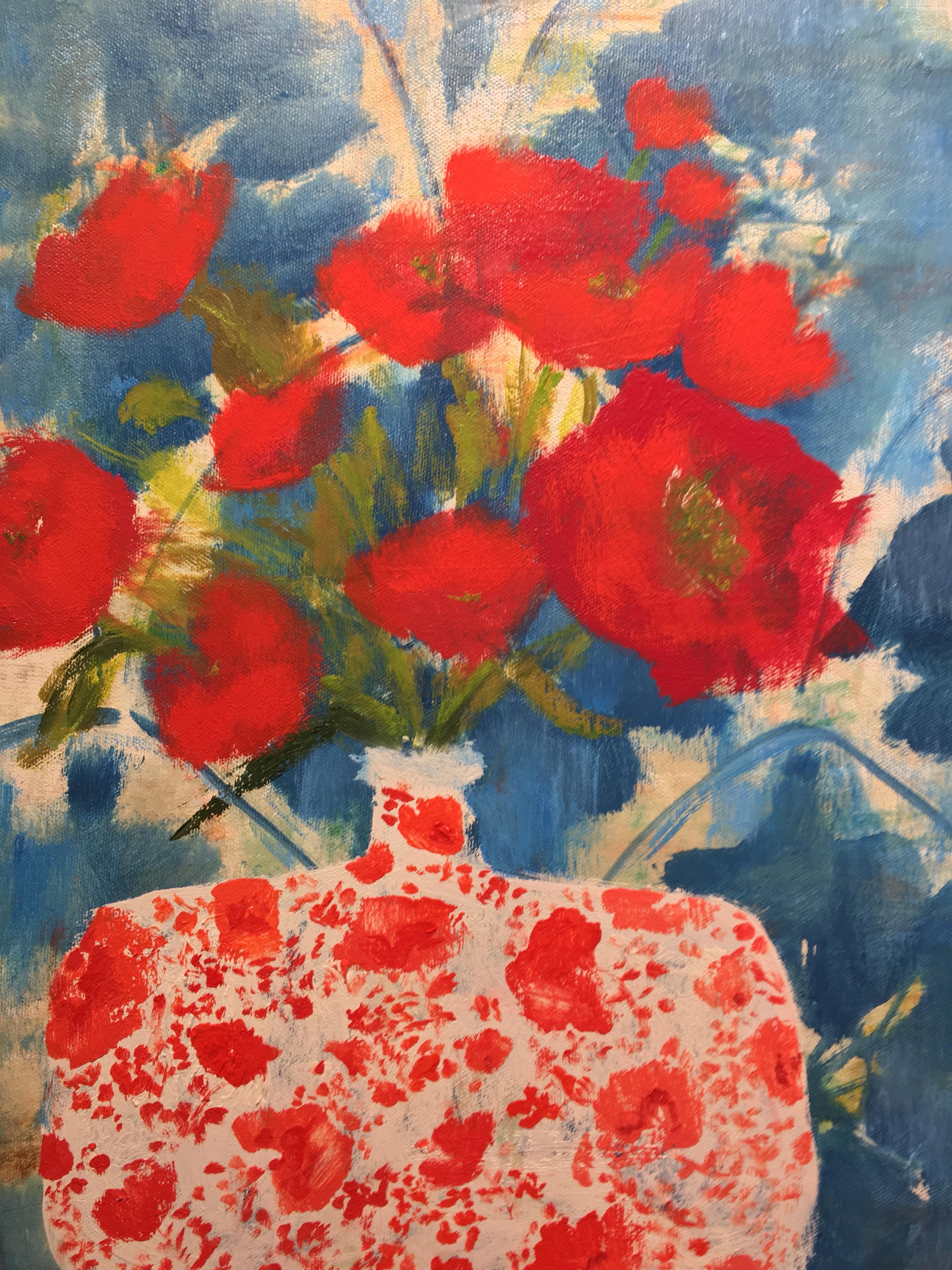 The bright red blossoms with green leaves in the bouquet at the center of this still life painting of flowers arranged in a red and white floral painted vase are beautifully complemented by the blue and white patterned wallpaper in the background.