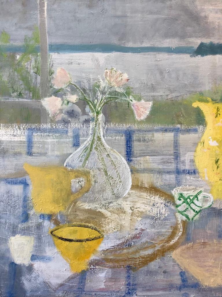 A contemporary painting in oil on canvas of a light and inviting interior scene with an arrangement of pale pink flowers in a clear glass vase next to canary yellow and green and white painted tea cups on a blue and white tablecloth. A pitcher with