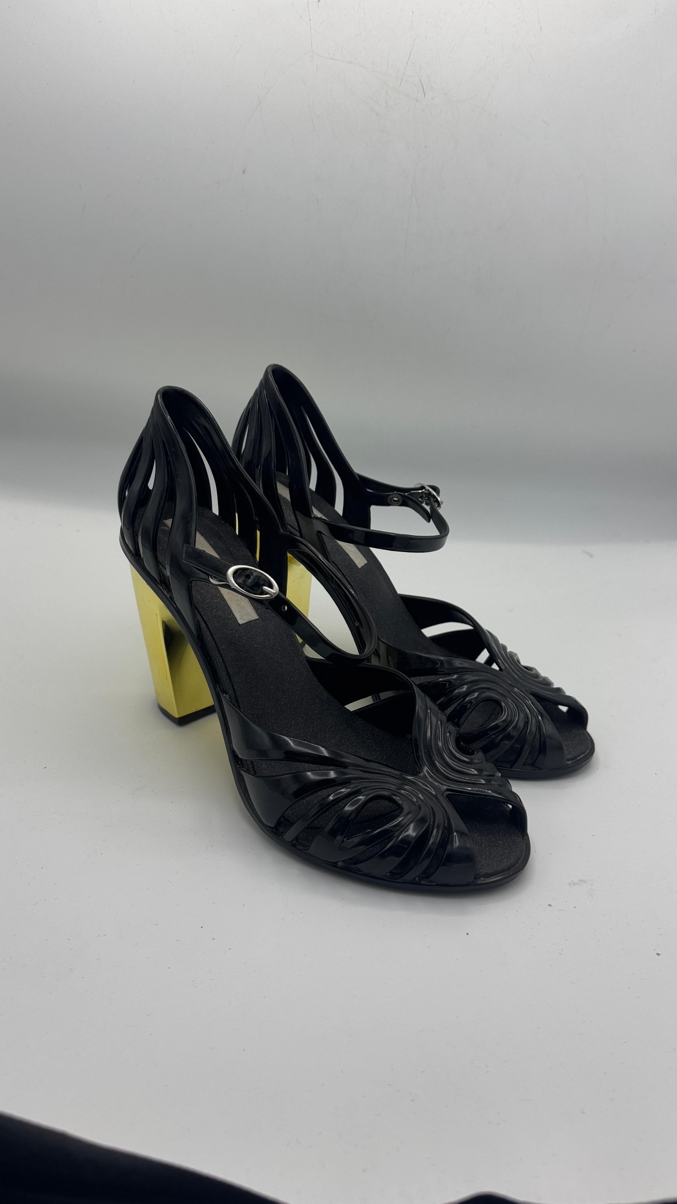 Striking and highly collectible vintage pair of Melissa heels crafted in glossy black PVC, featuring an intricate cut-out woven design and a dramatic gold geometric block heel. This bold silhouette is a testament to Melissa’s pioneering spirit in