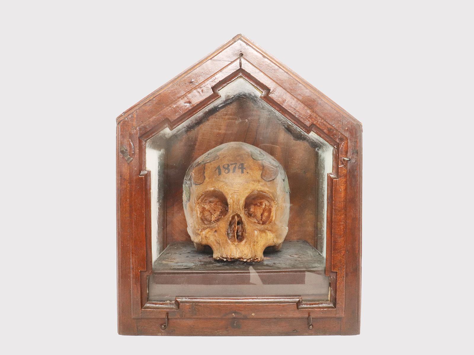 A Memento Mori from Wunderkammer, depicting a skull, plaster cast covered with butterfly wings, applied to the crown-shaped skull and to the mosaic-type wooden base. The year 1874 is painted on the forehead of the skull. Everything is kept inside a