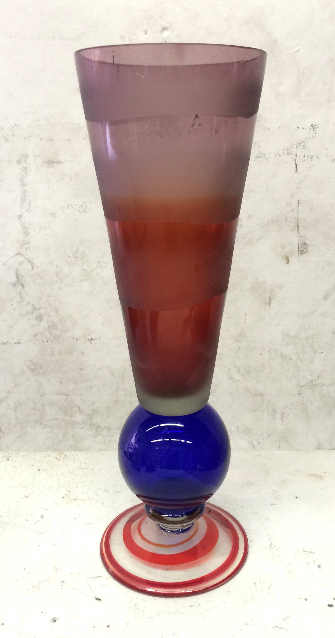 A fun Memphis Group-inspired colorful glass vase with contrasting shapes that seem to be stacked. The wide circular base is clear glass with swirling orange stripes, surmounted by a cobalt sphere that supports a purple and red semi-frosted