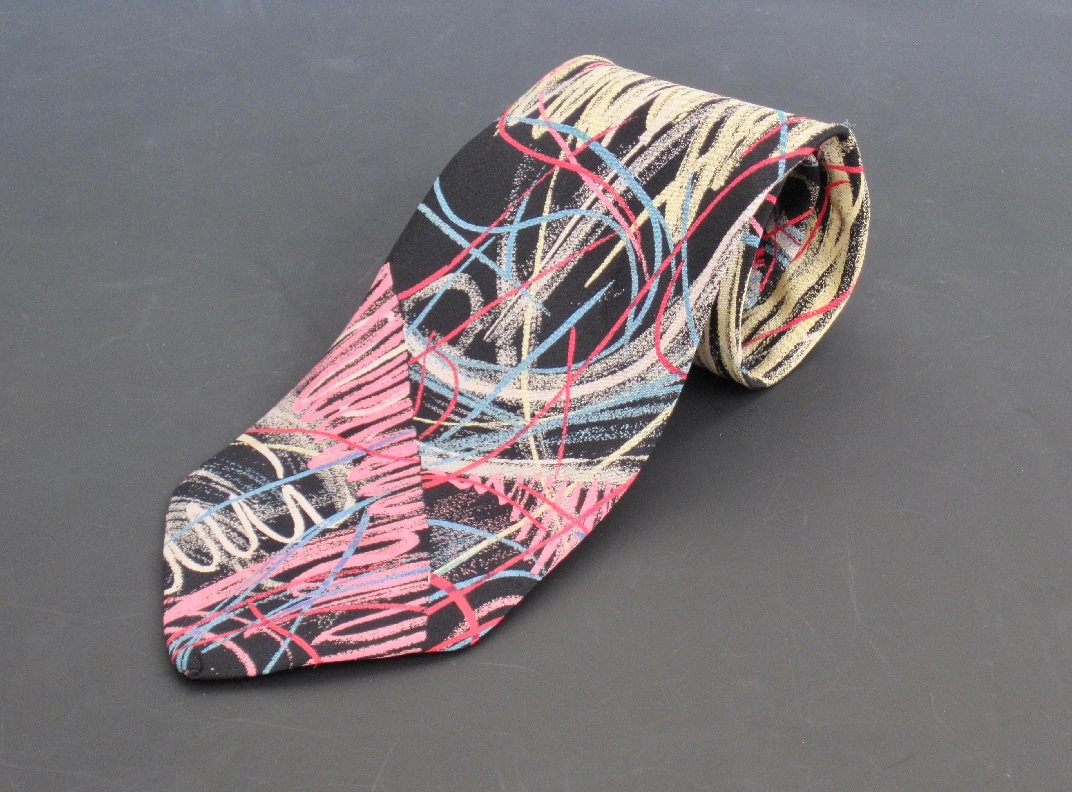 An Italian silk necktie with a colorful scribble-like design by Massimo Iosa Ghini, circa 1985. The blue and red line design over top a yellow, pink, and gray scribble print and black background create a fun, energetic, artful feel. 
This item has