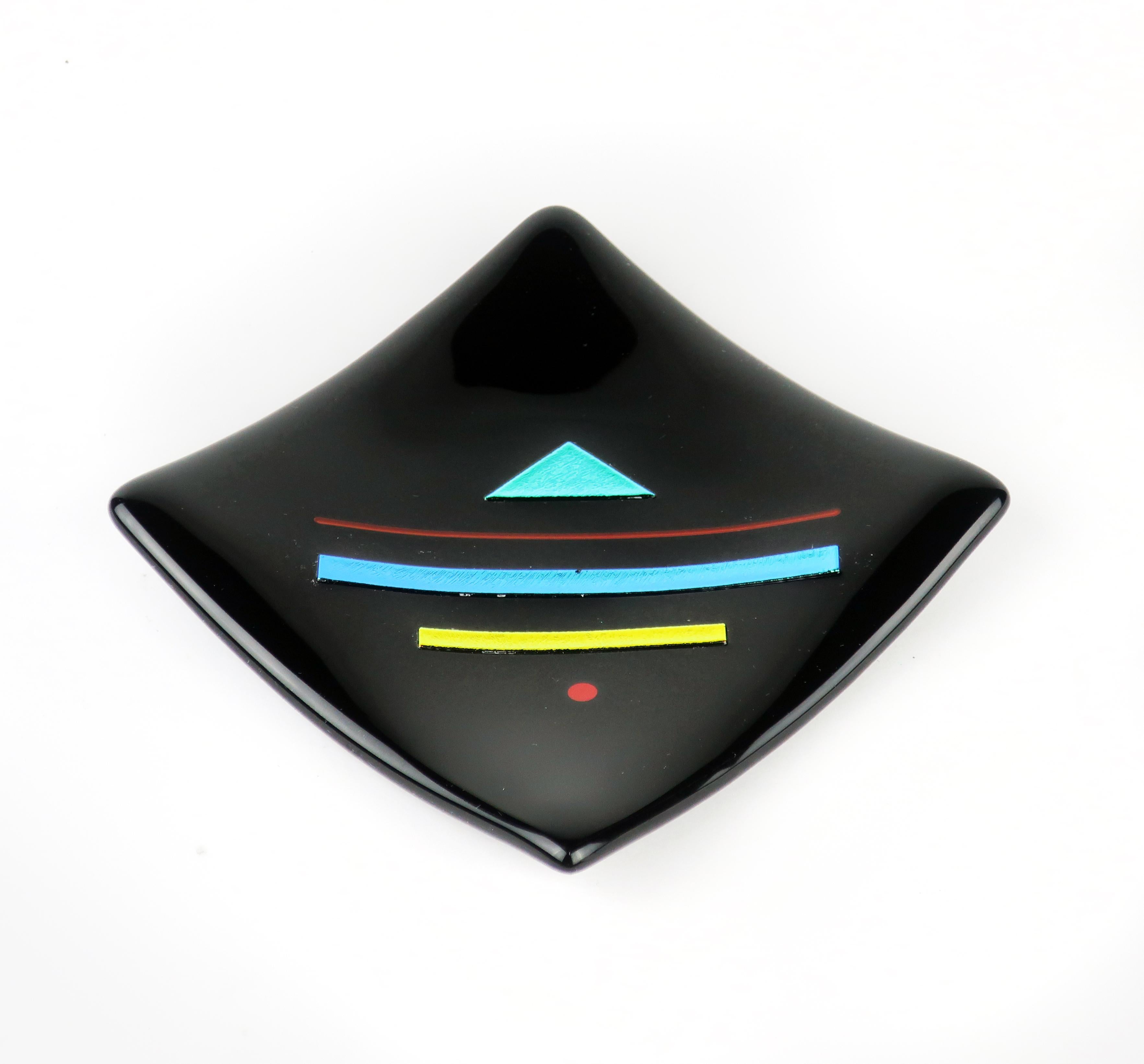 Strongly Memphis-influenced, this art glass dish by Karen Carlson has a black base with strips of rich blue, yellow and red glass that shift colors depending on the light.

Signed “Karen Carlson 94”.