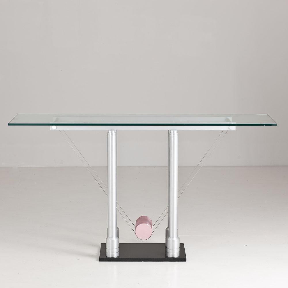 A brushed steel console table, set on a black steel base with a blush pink steel cylinder suspended by steel wiring. The console supports a glass top and is in the style of Memphis due to its quirky attribute, 1980s.