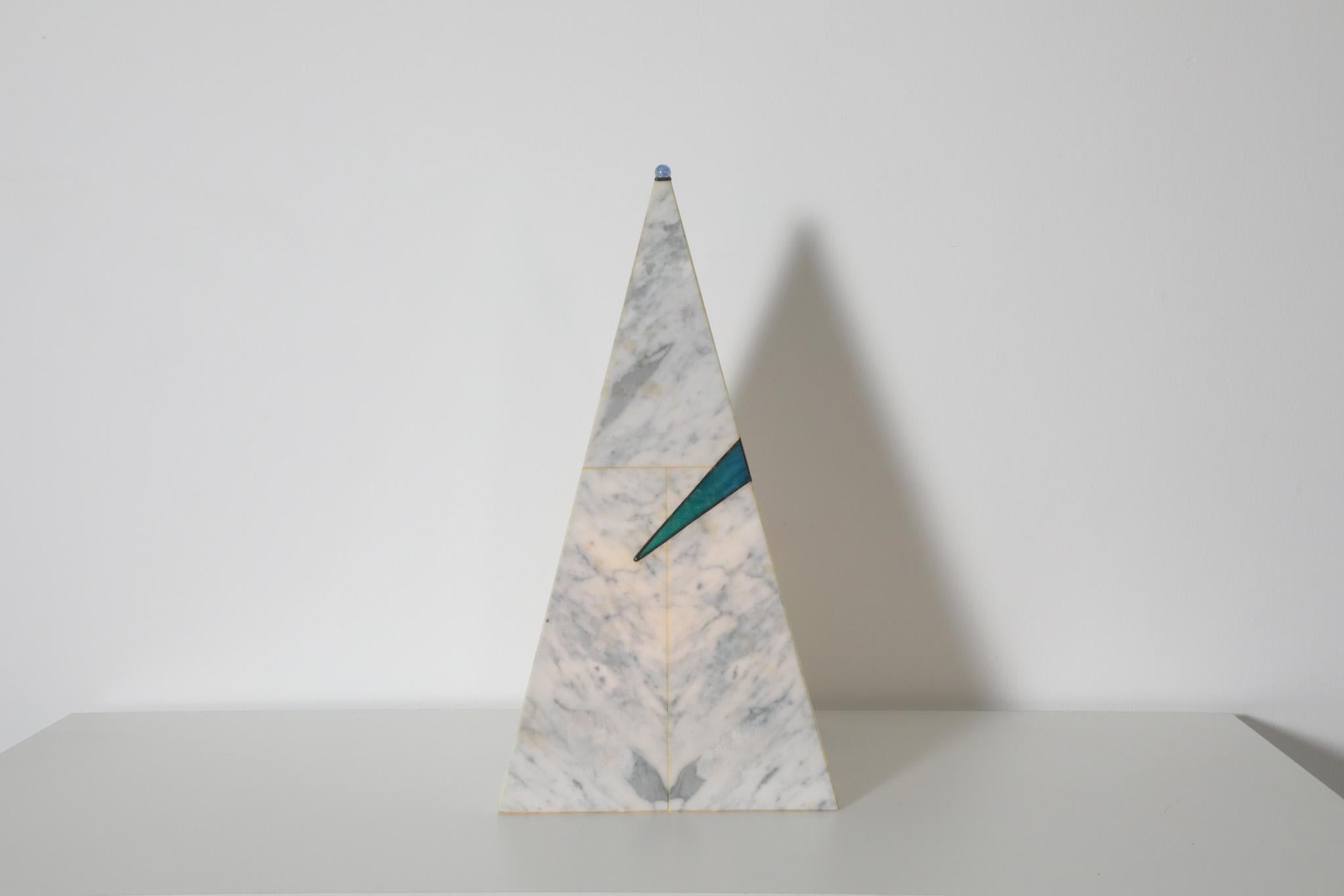 Pyramidenförmige Tischlampe im Memphis-Stil aus weißem und grauem Kunstmarmor mit einer kleinen Glasmurmel an der Spitze der Pyramide und einem blauen Kunstglasdetail in der Mitte. Die Lampe gibt einen schwachen Lichtschein ab. Im Originalzustand