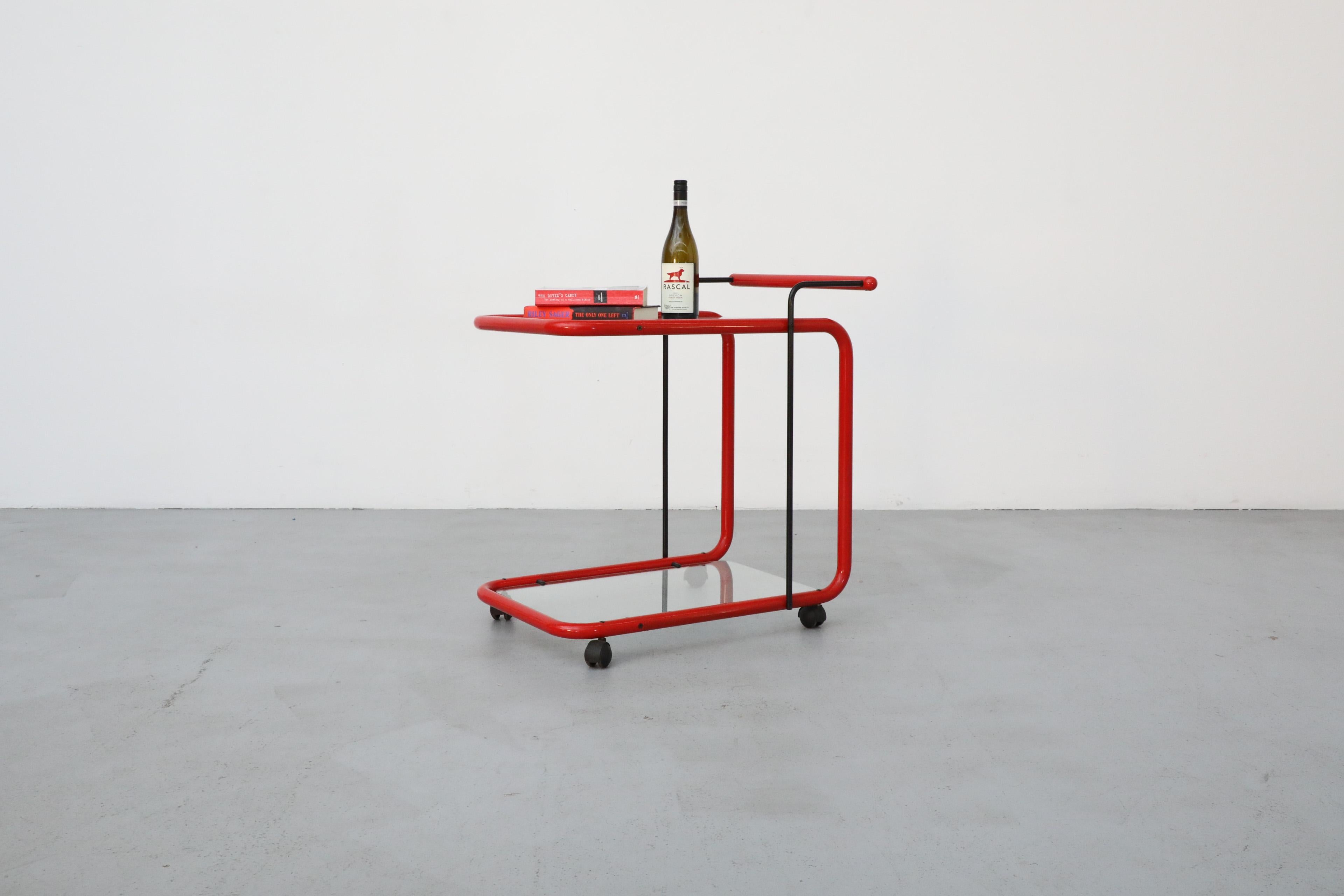 Memphis style post modern rolling bar cart with red tubular frame and two glass shelves. The top shelf has a white grid pattern on glass and the bottom shelf has been replaced with new glass. In otherwise original condition with visible wear