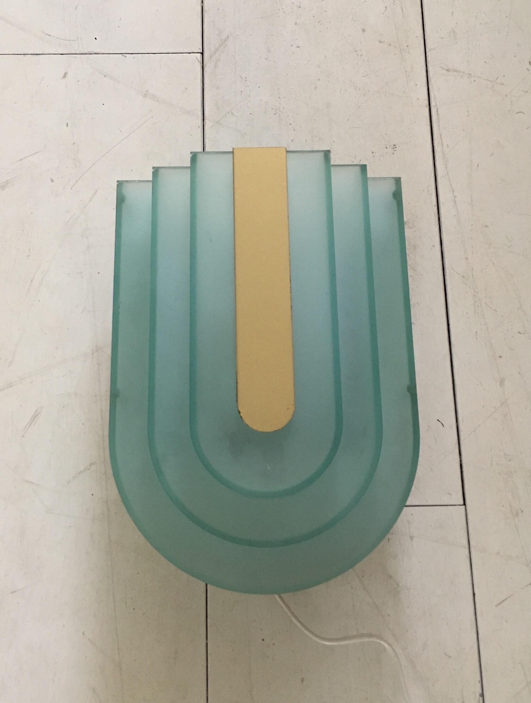 Typical piece from the 80's and very much influenced by Memphis, is this plastic and metal, aqua colored wall lamp. The plastic shade features a plastic mirror piece on top. The lamp remains in very nice condition with minor wear. It's signed