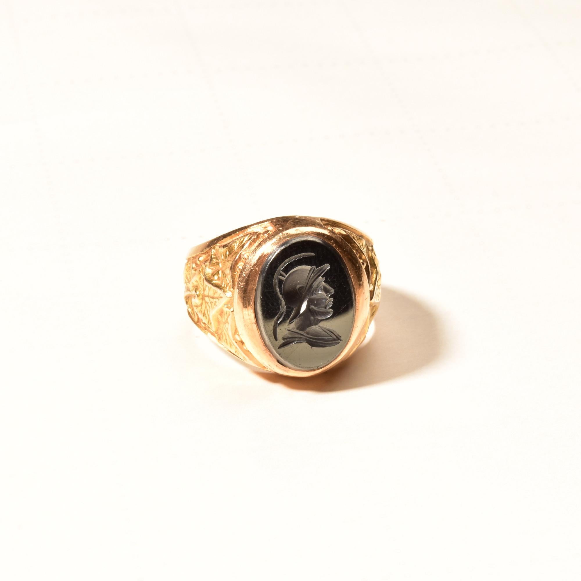 A timeless men's 18k hematite intaglio signet ring for a mid-century jewelry collection. Features a reflective gladiator cameo on a robust yellow gold band with raised leaf motifs. The ring is made from solid gold and has a considerable weight and