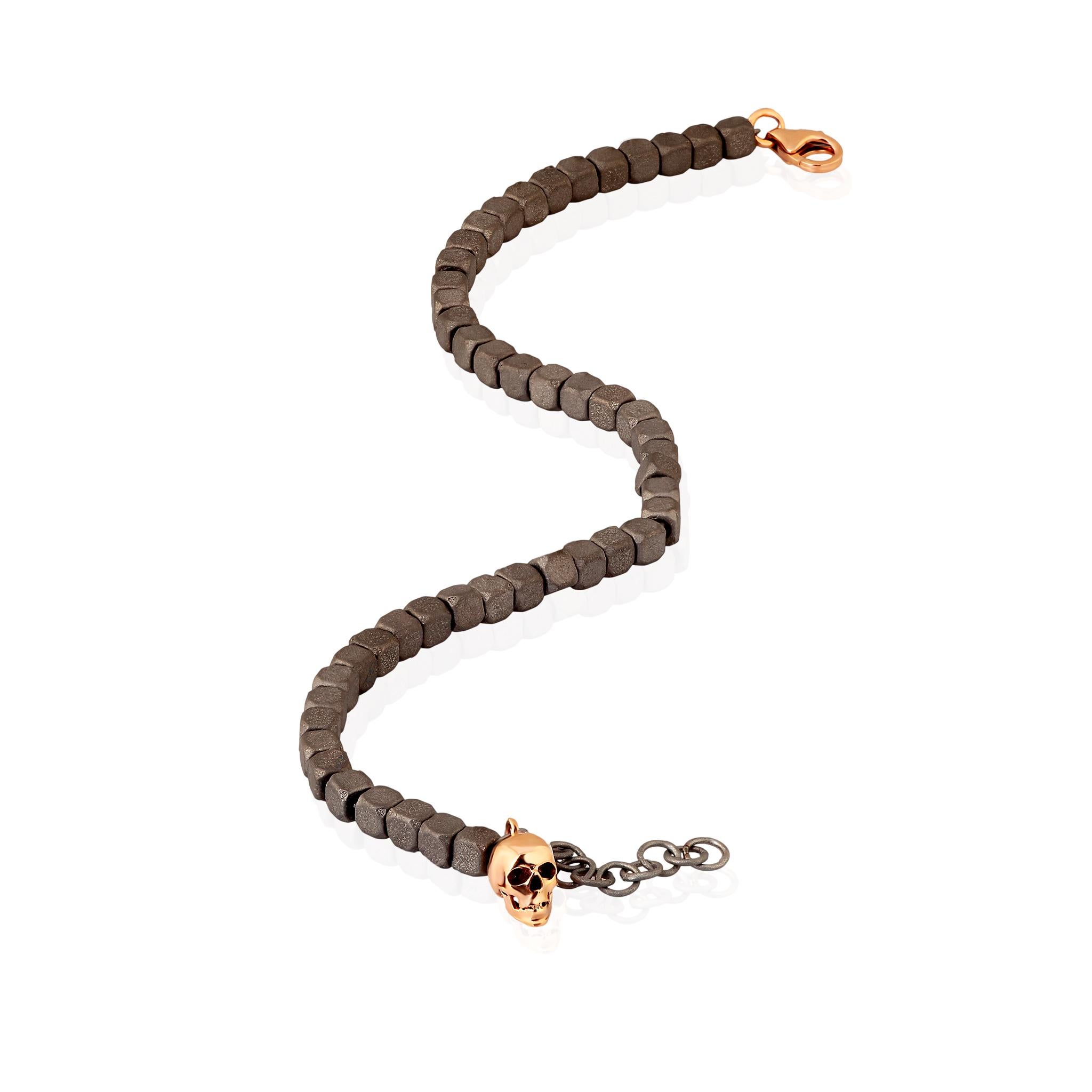 Bracelet en titane pour homme et pendentif tête de mort en or rouge 9 KT. Une série de petits cubes en titane constitue la structure principale du bracelet. Une chaîne traverse toute la section des cubes pour que le bracelet soit solide et mobile.