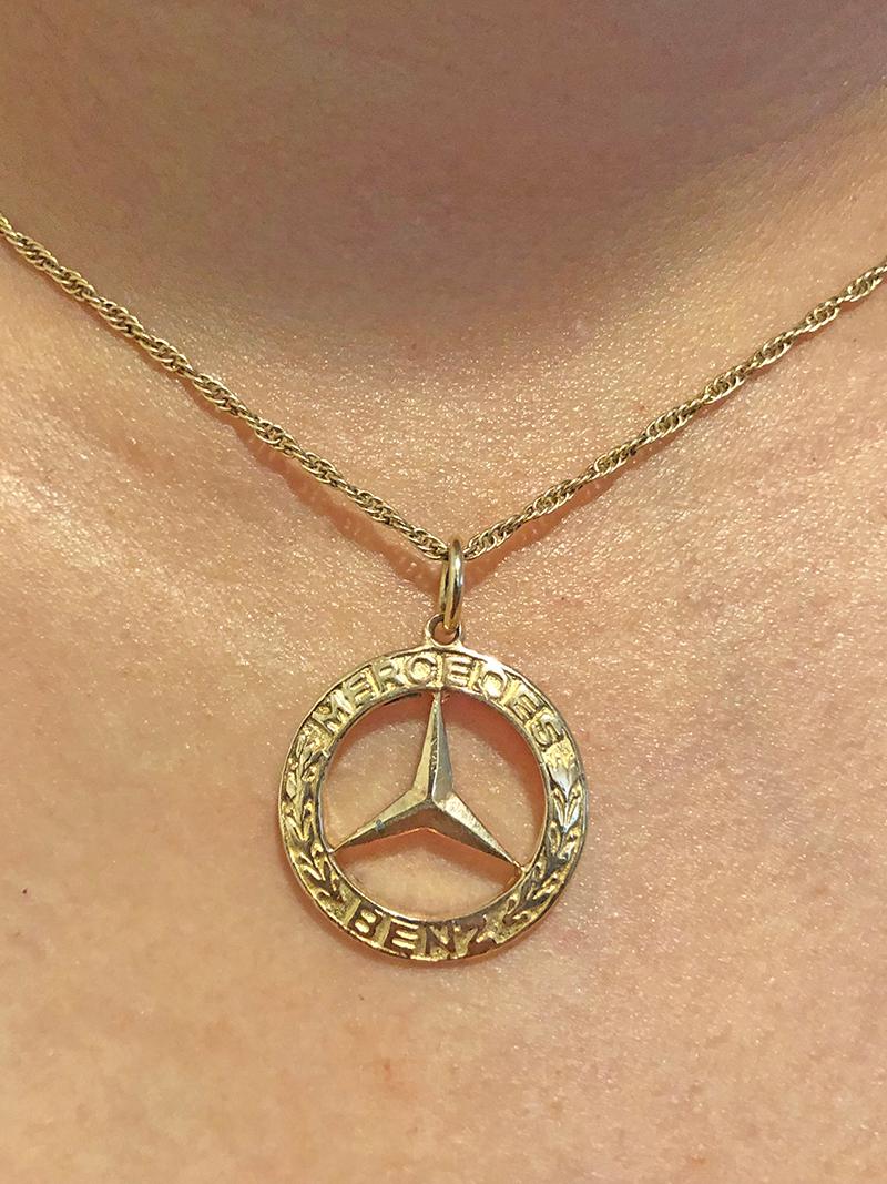 Round charm/pendant with cut-out Mercedes Benz logo in the center.  Applied letters around the border spell out 