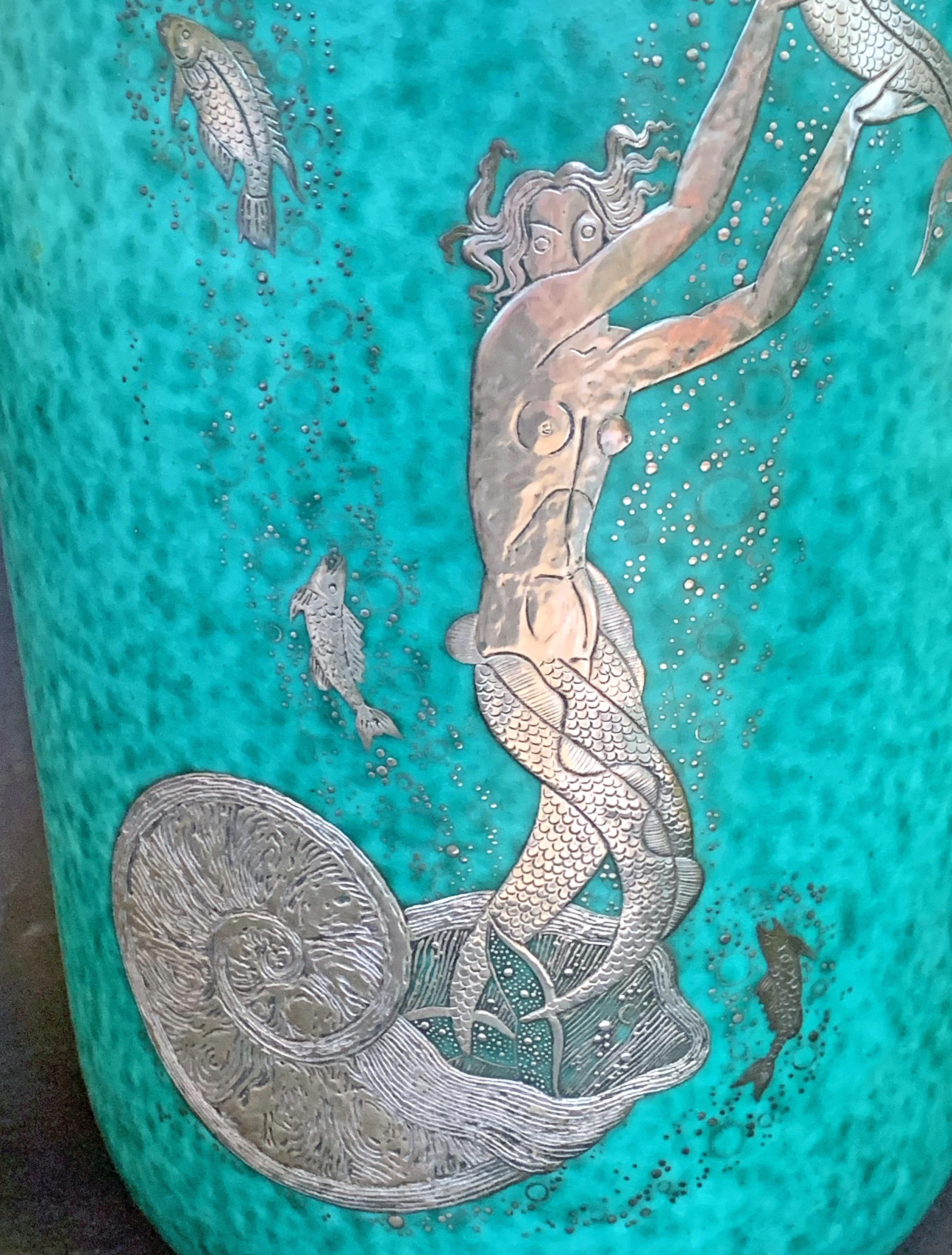Finished in tooled silver and a brilliant green-turquoise glaze, this unusually tall Argenta vase by Gustavsberg depicts a mermaid emerging from a nautilus shell, reaching for a large fish surrounded by a cloud of bubbles. This piece is part of a