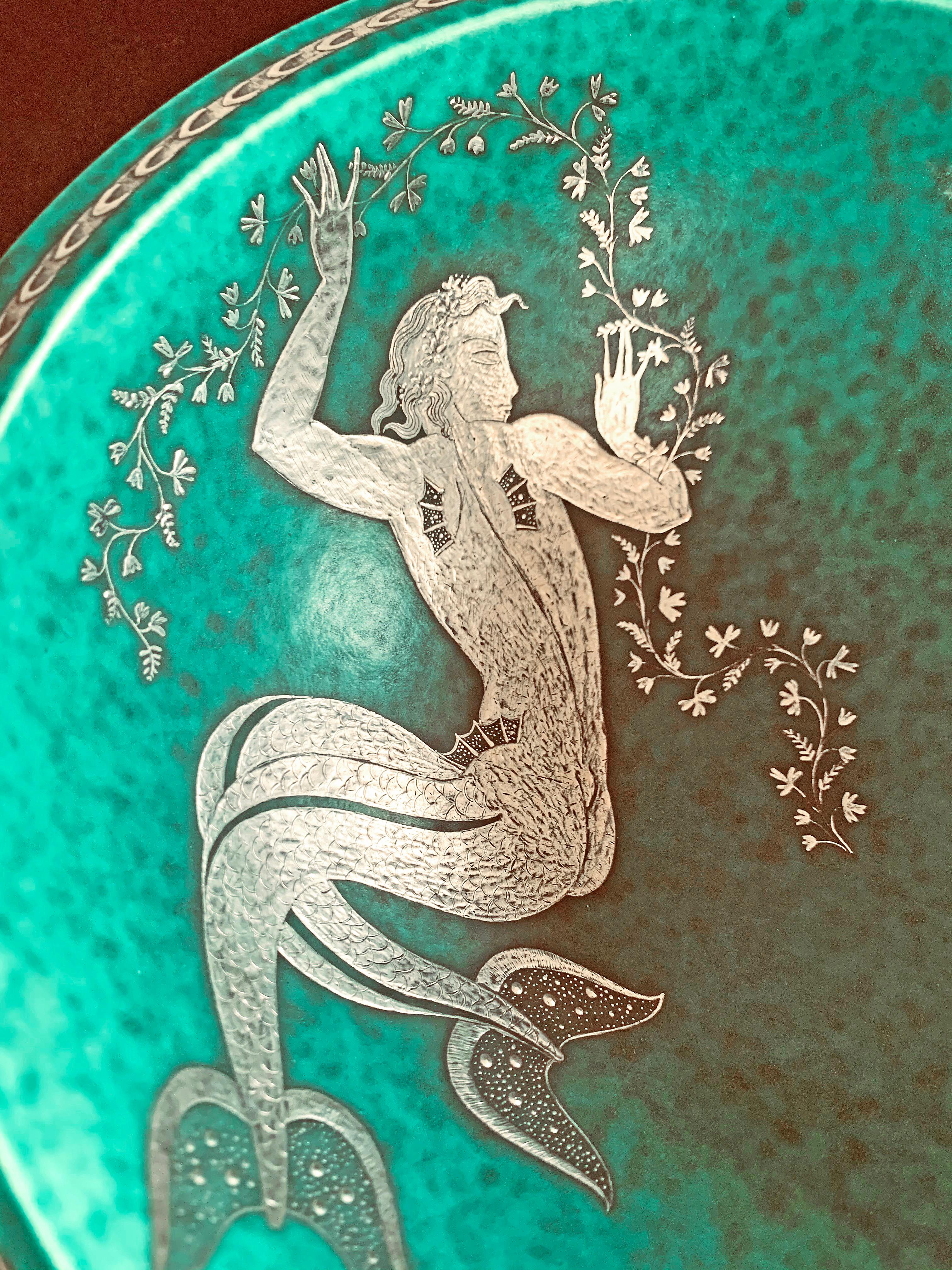 Finished in tooled silver and a brilliant green-turquoise glaze, this heavy porcelain bowl in the Argenta series by Gustavsberg is unusual in that its primary feature, a mermaid swimming in the sea while holding a flowered garland, has been applied
