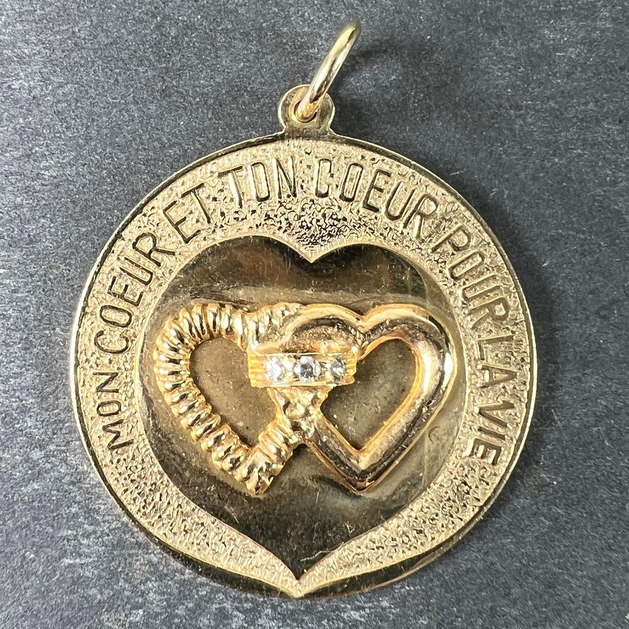Colgante amuleto del amor de oro amarillo de 14 quilates (14K) que representa un par de corazones, uno texturizado y el otro liso, unidos por una alianza engastada con tres diamantes. Los corazones tienen forma de corazón pulido sobre un fondo