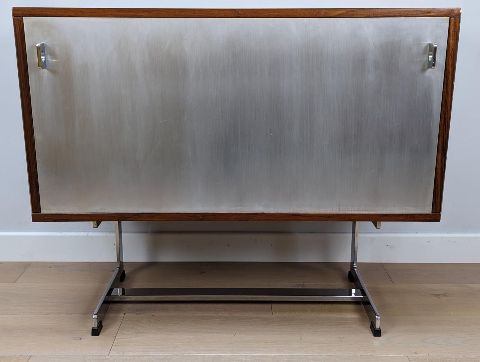 A stunning compact and extremely rare Merrow Associates sideboard / credenza.

The credenza is made out of Brazilian rosewood that has been professionally French polished. It has a drop down front that has a brushed aluminium front.

The unit
