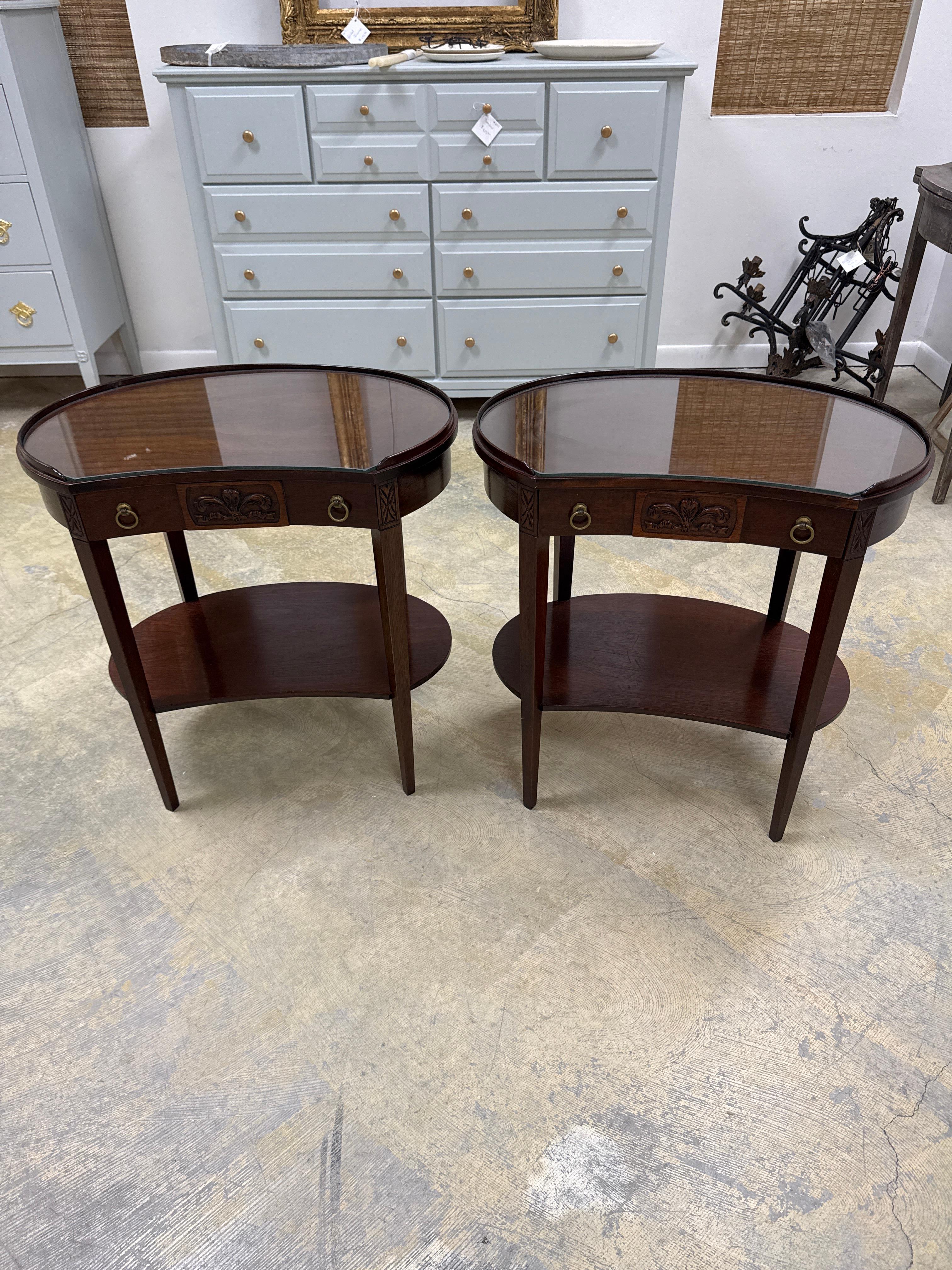 An antique mahogany Hepplewhite end tables with a glass top and a drawer. he two kidney-shaped tables in the image appear to be Mersman antique vanity console tables, likely made of mahogany wood with a glass top. Mersman tables were produced from