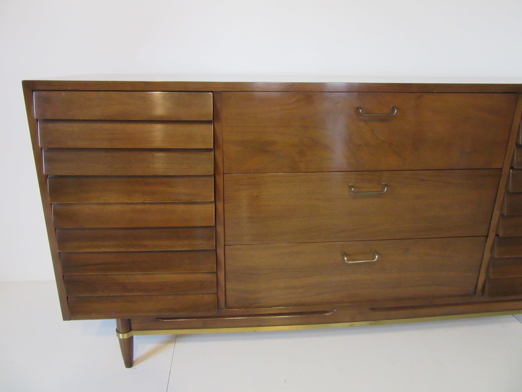 A dark walnut dresser with nine drawers, six with angled drawer fronts and three additional drawers behind two doors with brass pulls. Conical legs and wood stretchers with detailed brass to the lower leg area. Retains the manufactures branded mark,