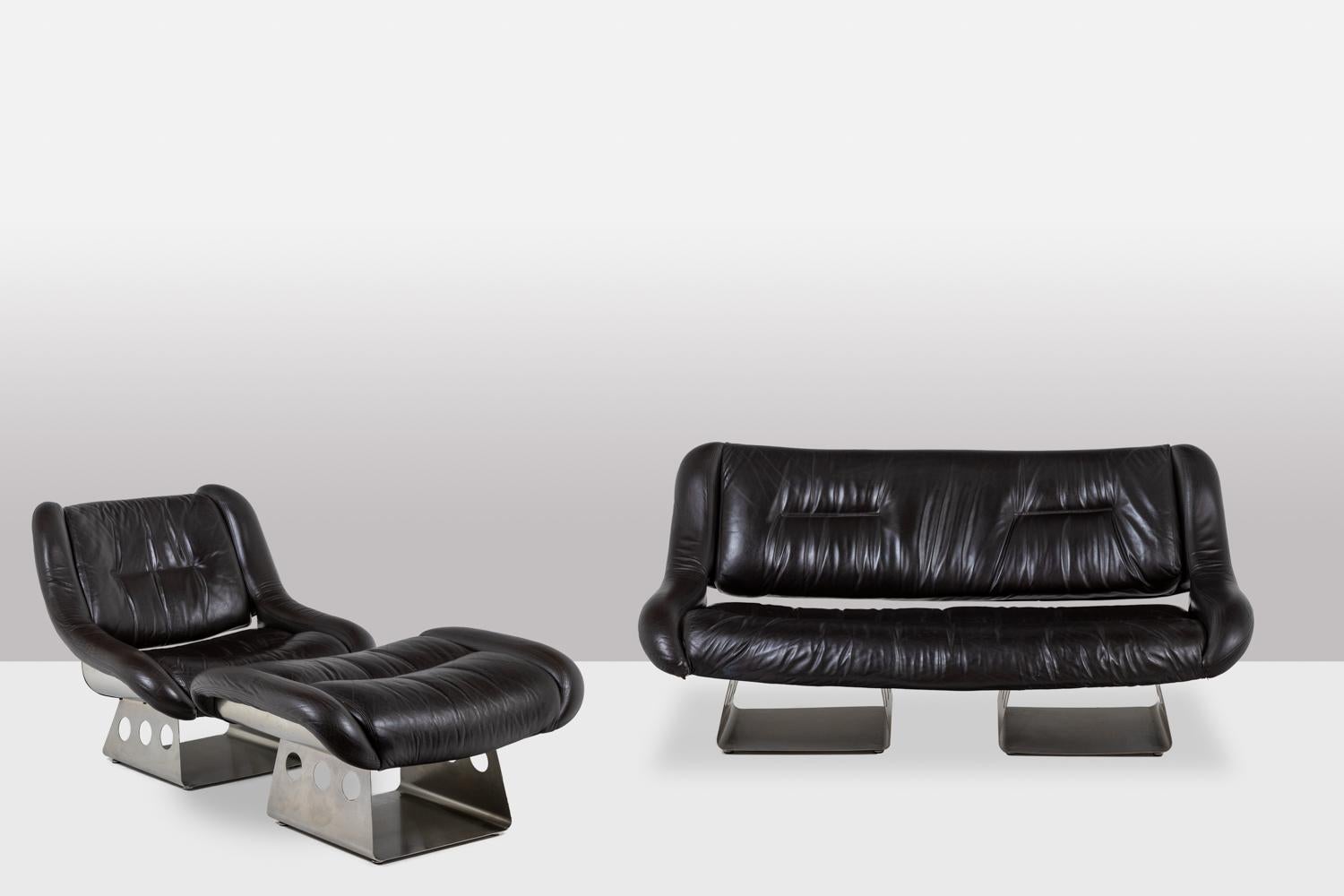 Wohnzimmer aus Metall und Leder, bestehend aus einem Sofa und einem Sessel mit Ottomane. Schokoladenfarbenes Leder. Sockel und Rückenlehne aus gebürstetem Edelstahl.

Das Werk wurde in den 1970er Jahren realisiert.

Abmessungen:

Sofa: H 80 x B 160