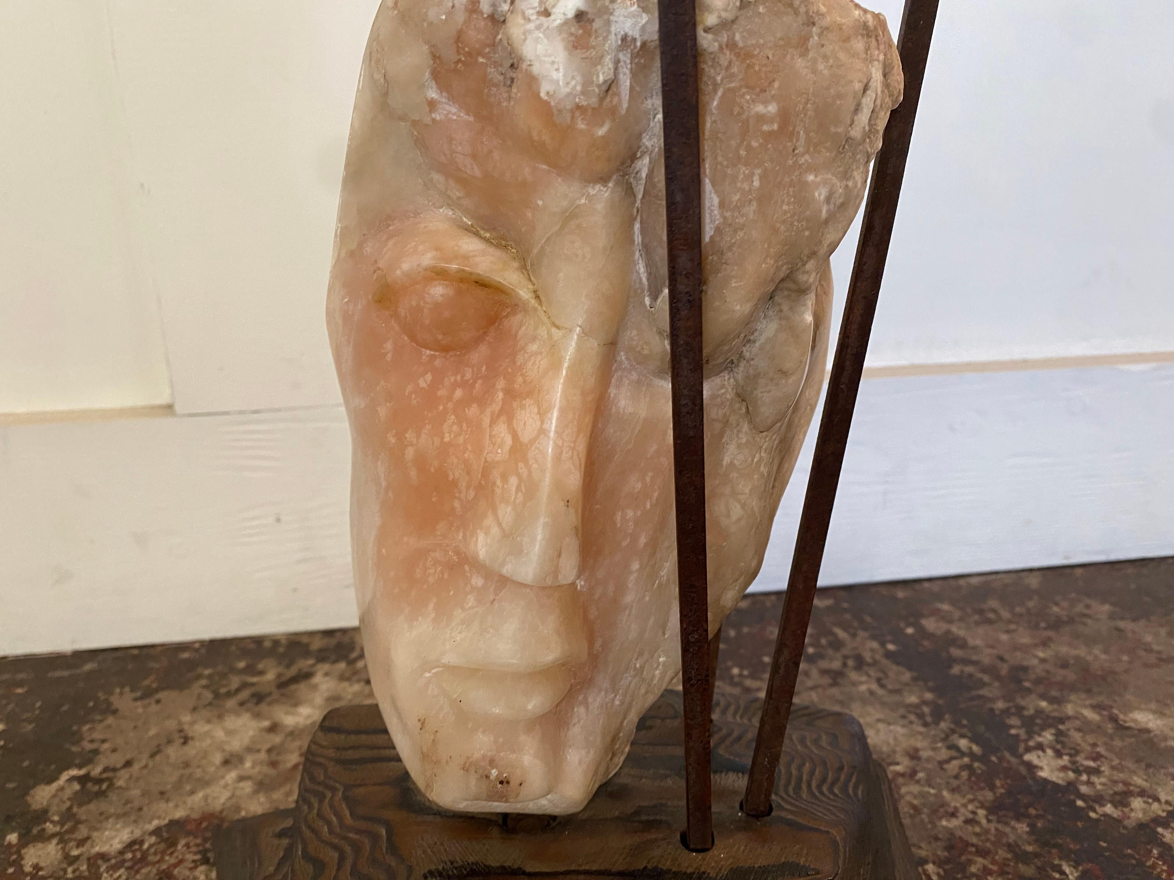 This marble sculpture, signed Leon Saulter, dated 1966, presents a stylized human face carved in light-toned stone, ranging from pale cream to soft pink. The coloration subtly recalls the natural tones of human skin.

The sculpture rests on a wooden