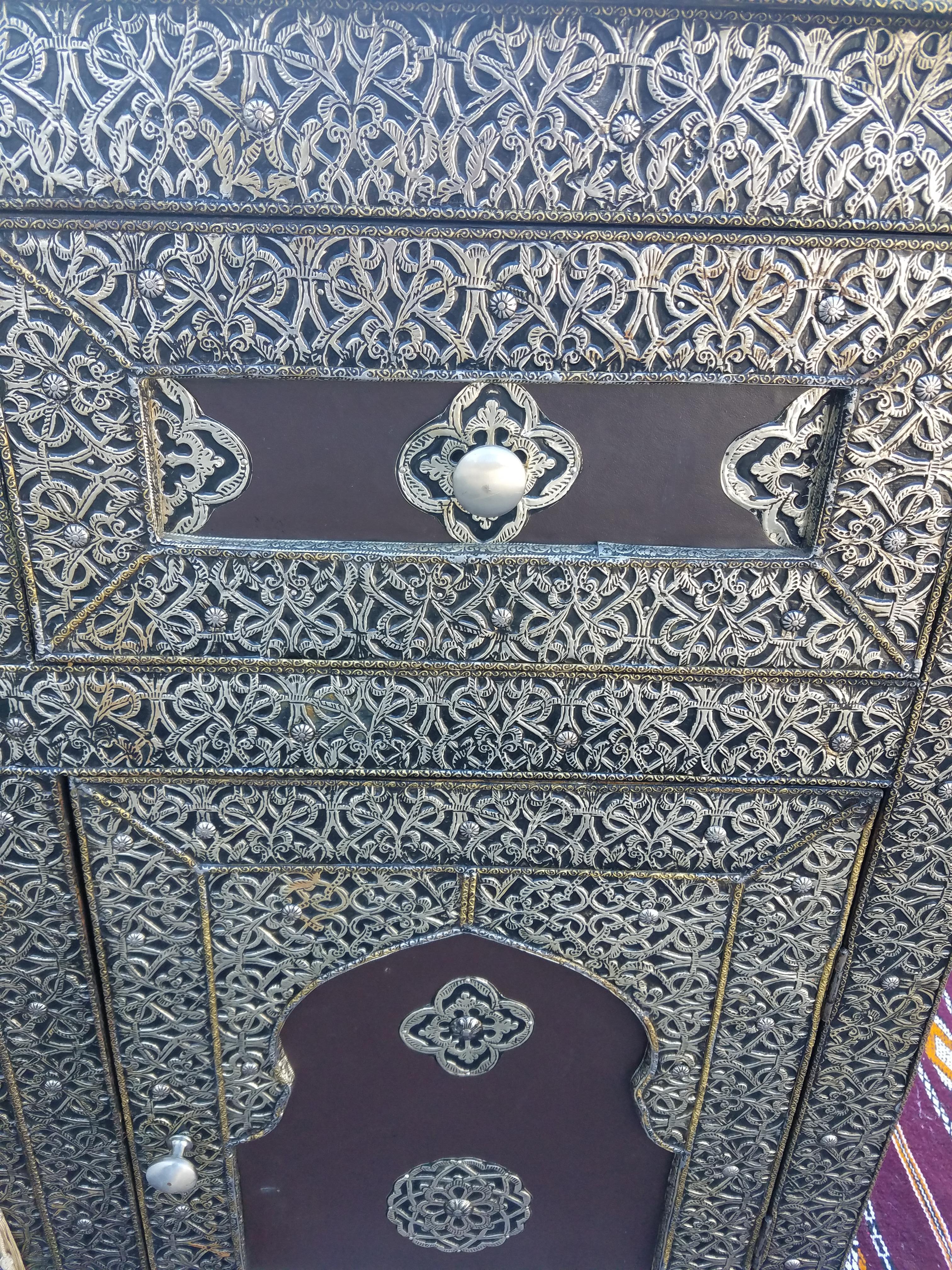 Aged all metal inlaid handmade Moroccan wooden cabinet measuring approximately 35” in height, 39” in width, and 16” in depth, and featuring one inside shelf with double doors and two easy to open drawers above. Plenty of storage. Lots of details