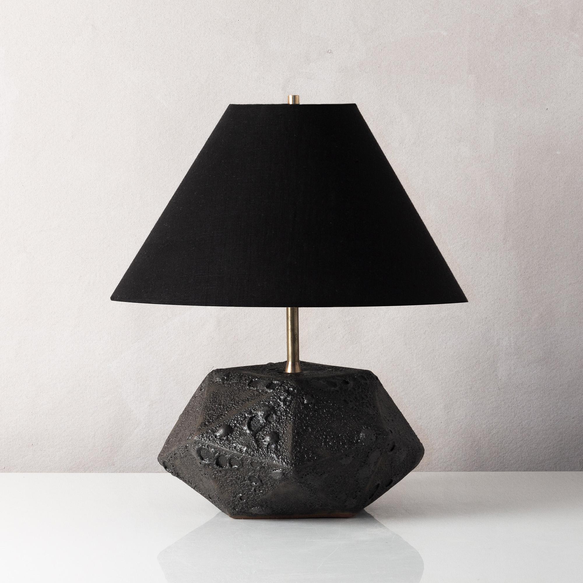 This dramatic black ceramic table lamp features a complex geometric shape, accentuated by a textured black glaze reminiscent of volcanic rock. Each piece is individually handmade and entirely unique. The lamp is finished with raw brass hardware and