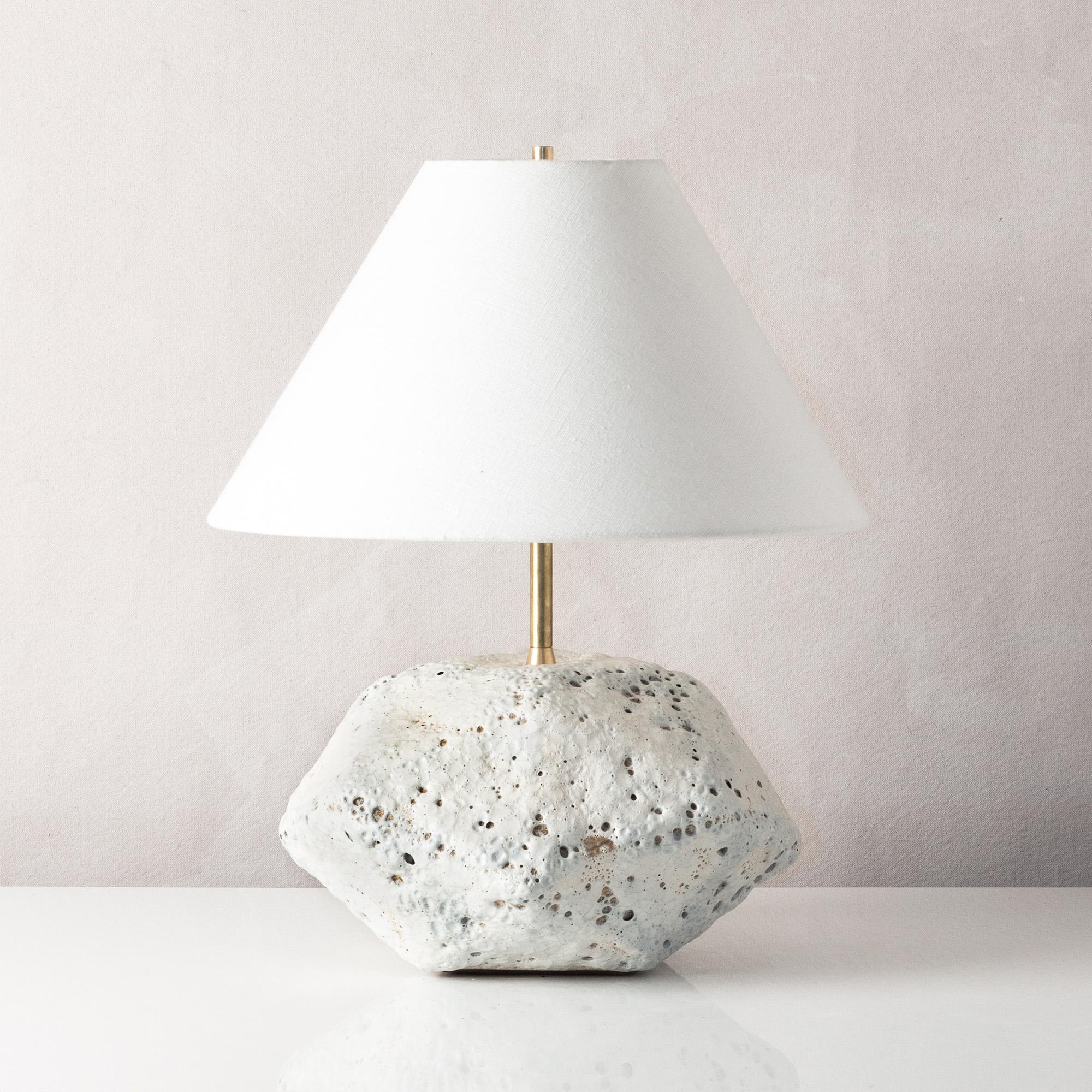 This dramatic white ceramic table lamp features a complex geometric shape, accentuated by a textured white glaze reminiscent of volcanic rock. Each piece is individually handmade and entirely unique. The lamp is finished with raw brass hardware and