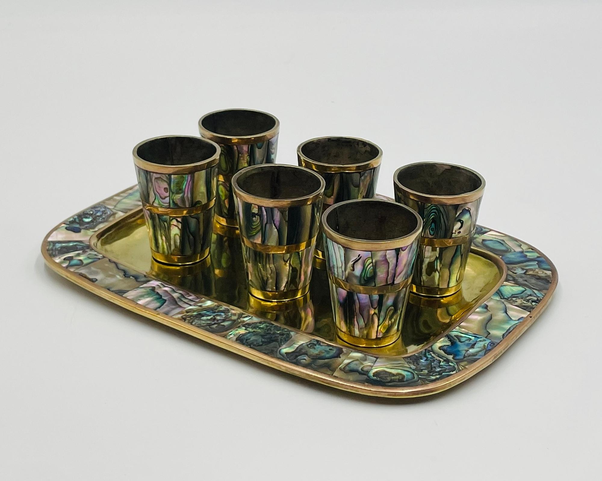 Striking mid-century Mexican shot glass and tray set, made of brass with inlaid abalone shell. The set includes six shot glasses and a matching rectangular serving tray, all showcasing the iridescent beauty of natural abalone in rich tones of green,