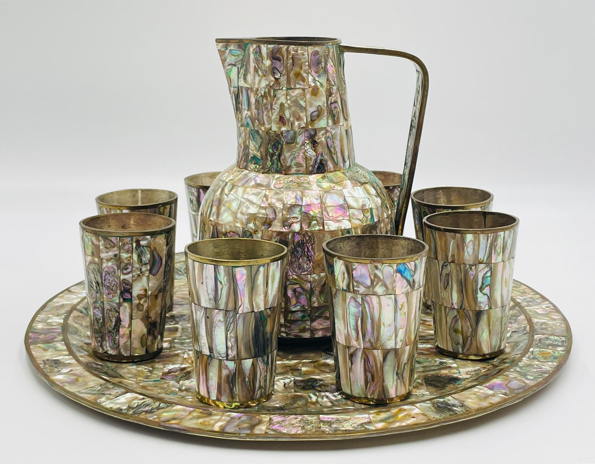 Elegante juego de servicio mexicano de mediados de siglo que incluye una bandeja redonda, una jarra de agua y ocho tazas a juego, fabricadas en latón macizo con exquisitas incrustaciones de concha de abulón. Cada pieza muestra la iridiscencia