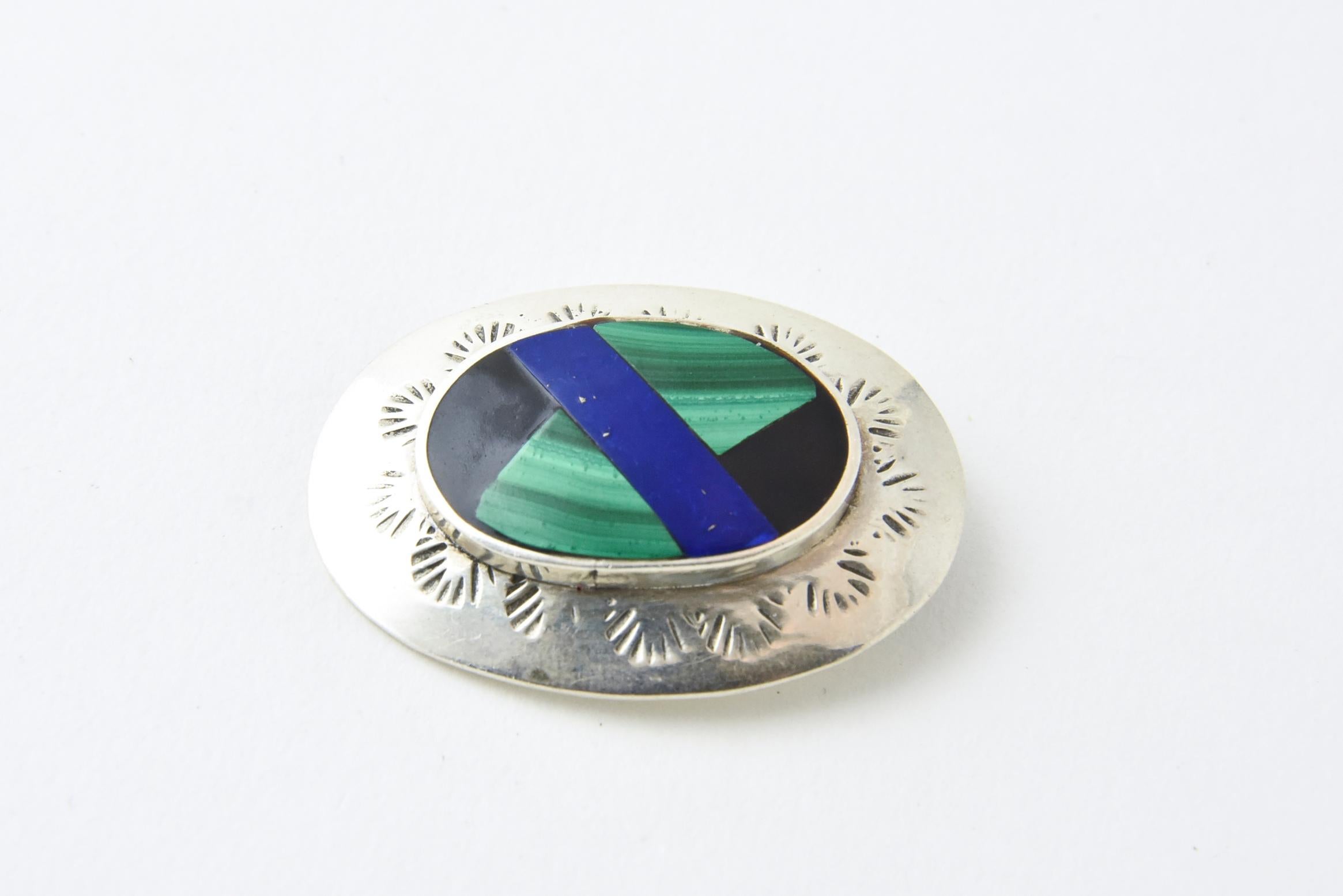 Black onyx, malachite and lapis inlaid into an oval sterling silver brooch. The raised inlaid section is surrounded by a frame with an etched sun design. Marked: Mexico 925.