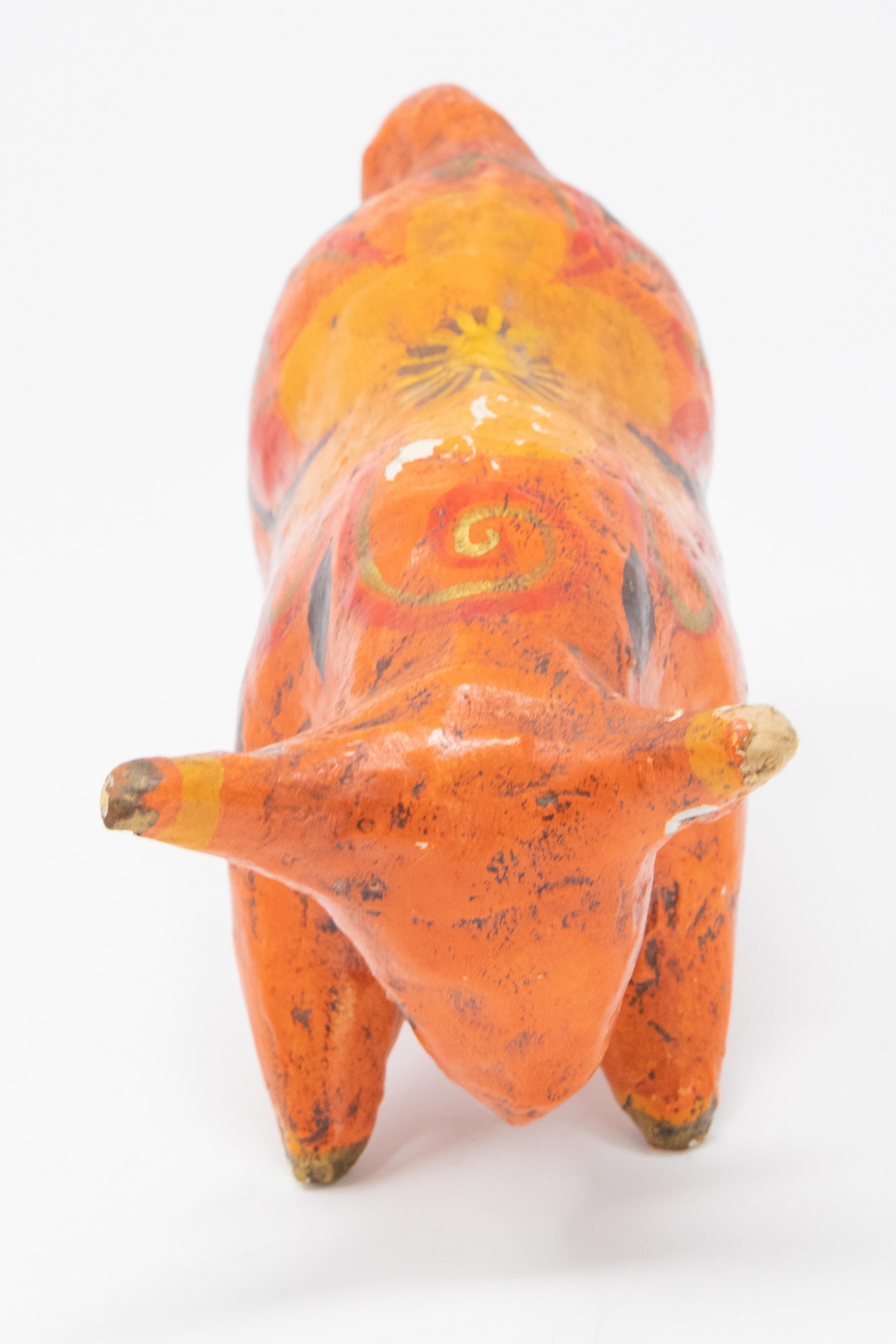 Featured is a Mexican bull figurine. This figurine has been painted a vibrant orange color with a black and yellow floral design on its back. This piece is marked “MEX” on the underside of the figurine. It is made of papier mâché.