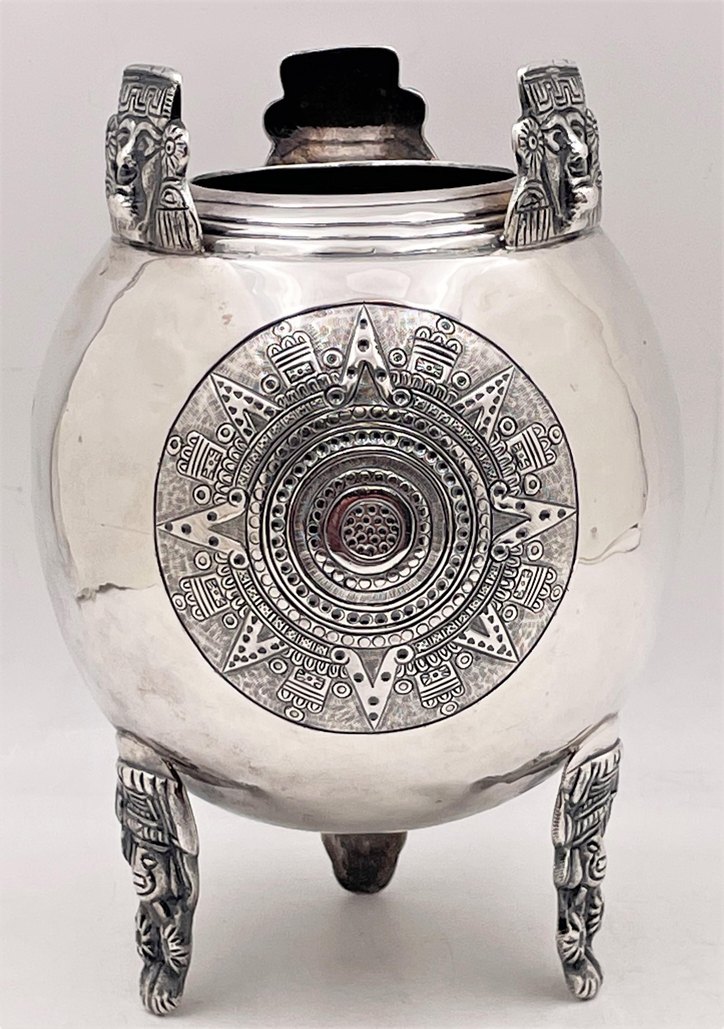 Vase mexicain en argent sterling, présentant des motifs aztèques élaborés, dont trois différents sur le corps, reposant sur trois pieds avec des têtes figuratives, ainsi que des masques de style aztèque émergeant du sommet du vase. Le poinçon est