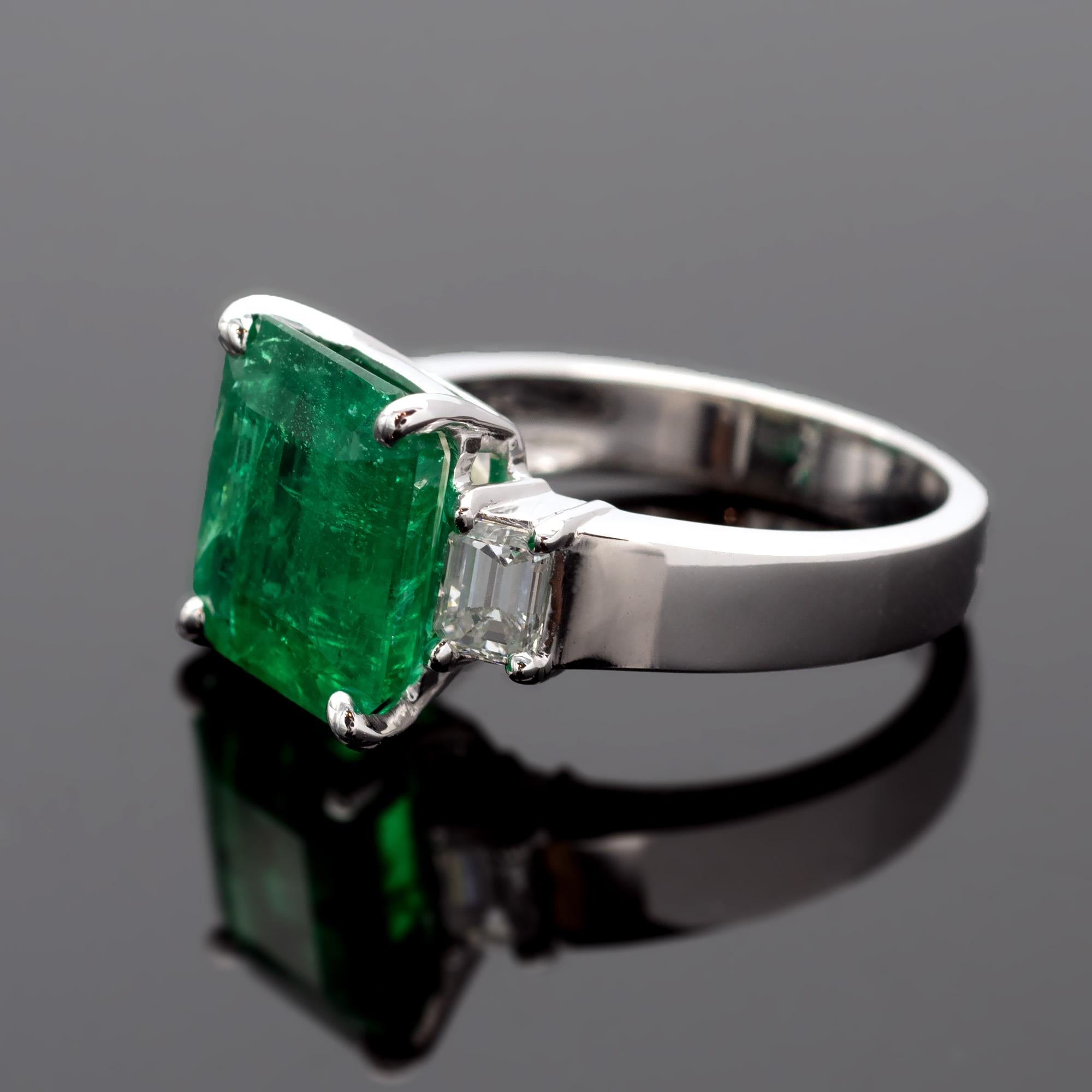 Vibrant coloured nearly 5 carat Colombian emerald set with two emerald cut diamonds weighing 0.58 carats  ( FG VS1 )
The ring itself is a very well made 18 Karat white gold engagement ring, modern yet timeless. 
The emerald comes with a gem report
