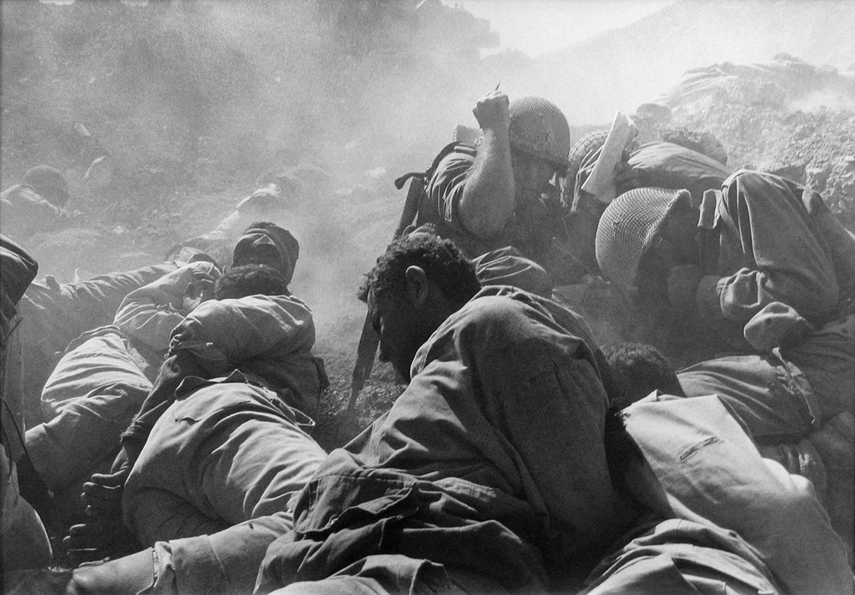 Artillery Barrage, Suez Canal, Yom Kippur War by Micha Bar-Am depicts soldiers amidst a battle. The men are laying on the ground, ducking for cover in the dusty atmosphere. Surrounded by rubble, the men brace for impact of the next shot of