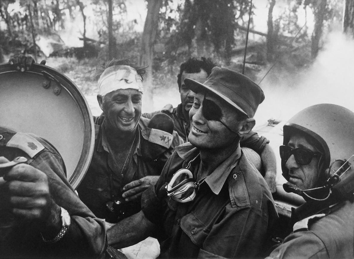Crossing the Suez Canal, Yom Kippur War by Micha Bar-Am presents a brief moment of joy amidst a dark time. Two men smile while riding in a car, with other men looking onward with a blank face. One man has a bandage wrapped around his head, while the