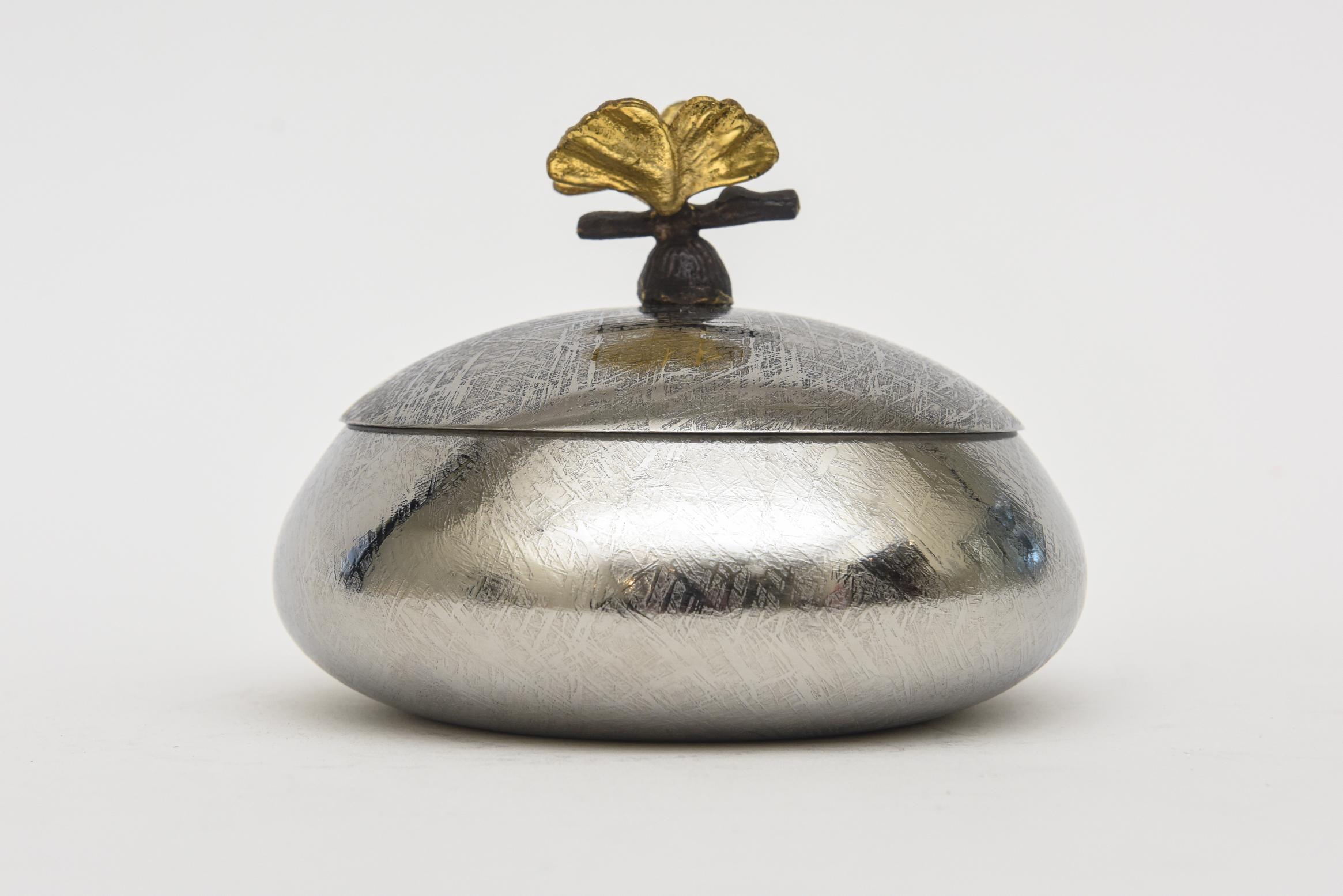 This lovely small round 2 part box is an early piece of the designs of MIchael Aram's work from the early 90's.  It is a tooled design silver metal on the top of the box with a blackened metal and brass flower top. The interior is a cream enamel