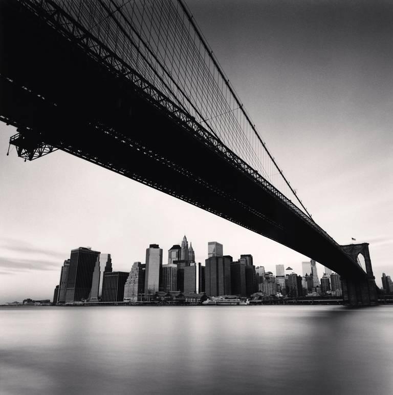 Brooklyn Bridge, Studie 1, New York, USA von Michael Kenna zeigt eine dramatische Szene. Die Brooklyn Bridge spannt sich in den Himmel und führt zur Skyline von New York City am Horizont. 

Auflage 30/45
Sepia getönter Gelatinesilberdruck
Bildgröße: