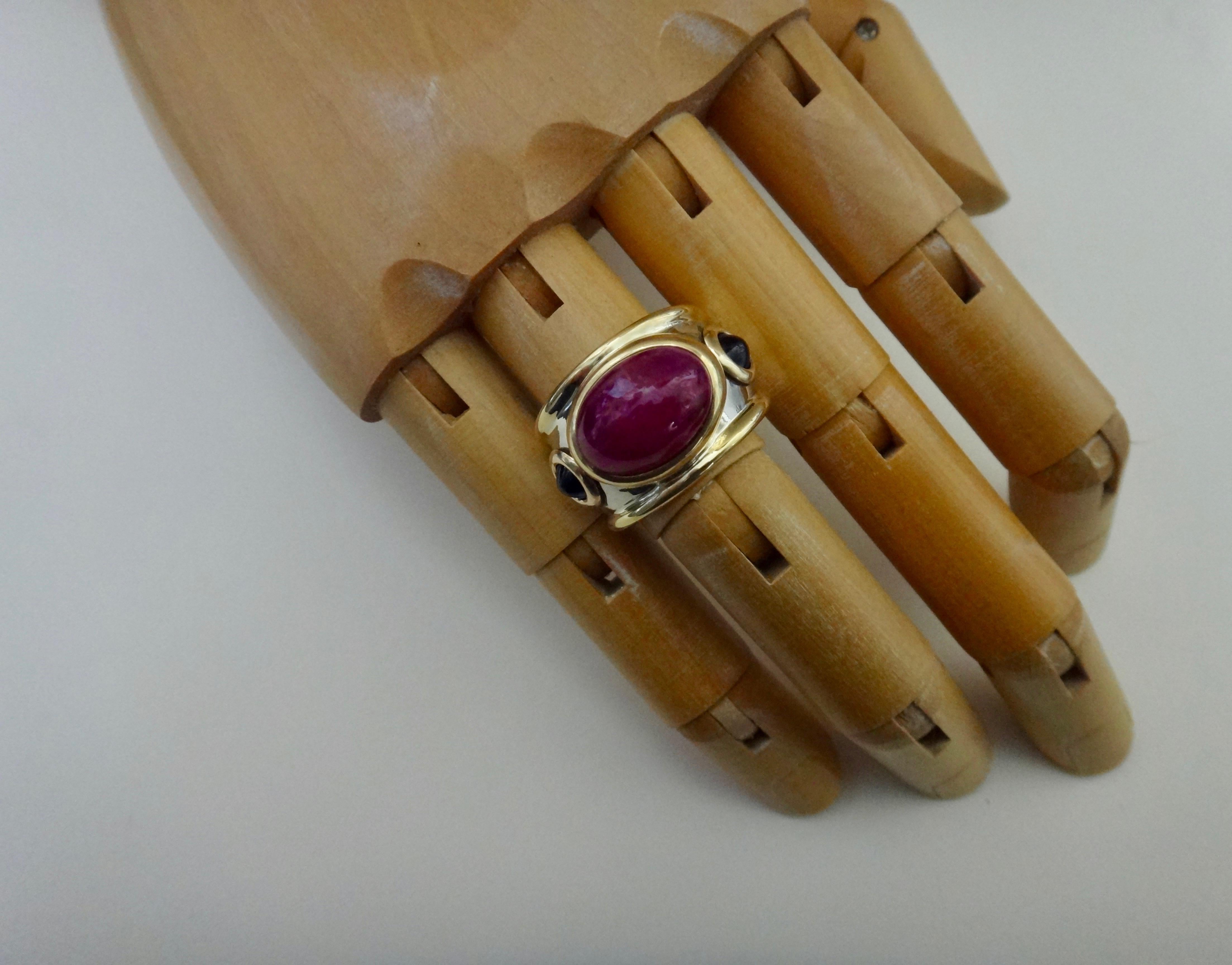 A high domed cabochon ruby (origin: Thailand) of a bright pinkish/red color is bezel set in an hand fabricated 18k yellow and white gold Bombe ring.  The gem is flanked by a pair of cabochon blue sapphires (origin: Thailand) of a rich blue color and