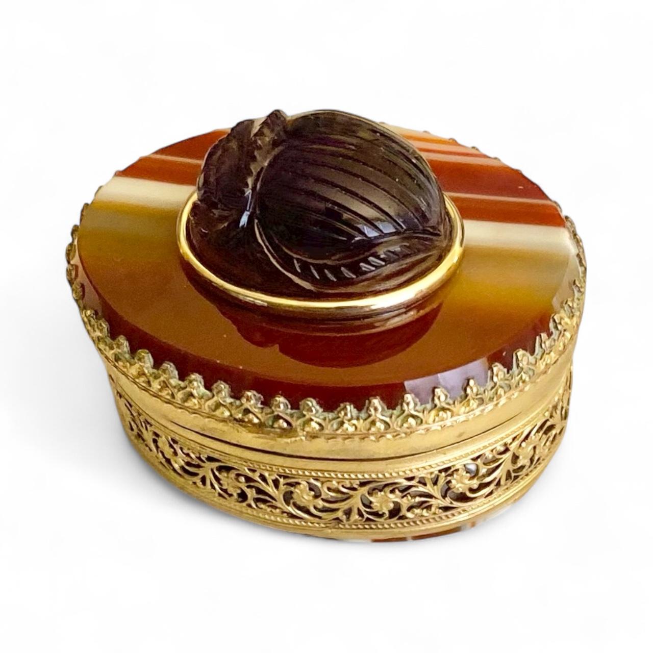A tiny Victorian box is embellished with an amethyst scarab.  This box, known as a vinaigrette, pomander or scent box was used to disseminate fragrance.  The box is elegantly crafted from banded agate, both top and bottom.  All set in a pierced gilt