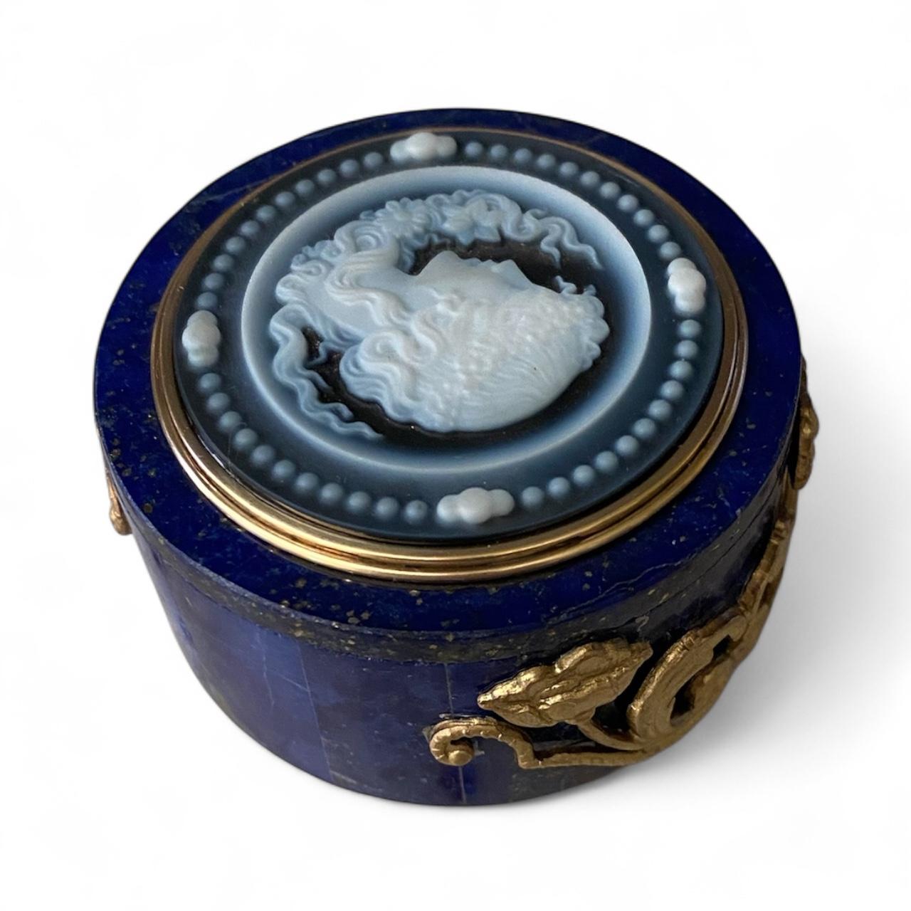 A hardstone cameo is featured in this distinctive ring box.  The cameo is carved from blue and white banded chalcedony in the form of a graceful young woman..  it is framed in gold and mounted on a box of lapis lazuli.  Decorating the body of the