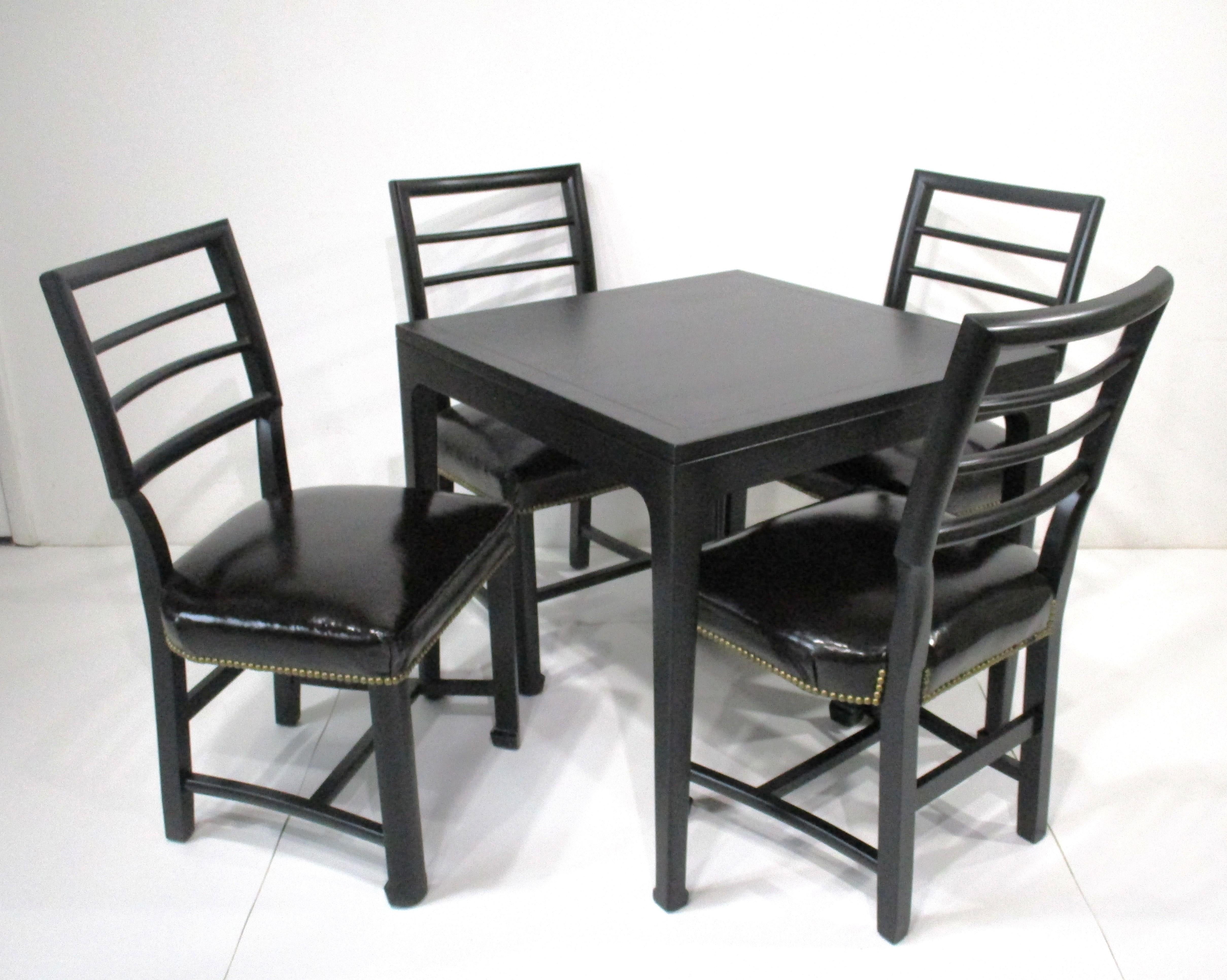 A very well crafted game or dining table set with four matching chairs all in a satin black finish . The ladder back chairs are upholstered in a luxurious soft patten leather with brass stud tacking to the edges . The tables finish has that semi