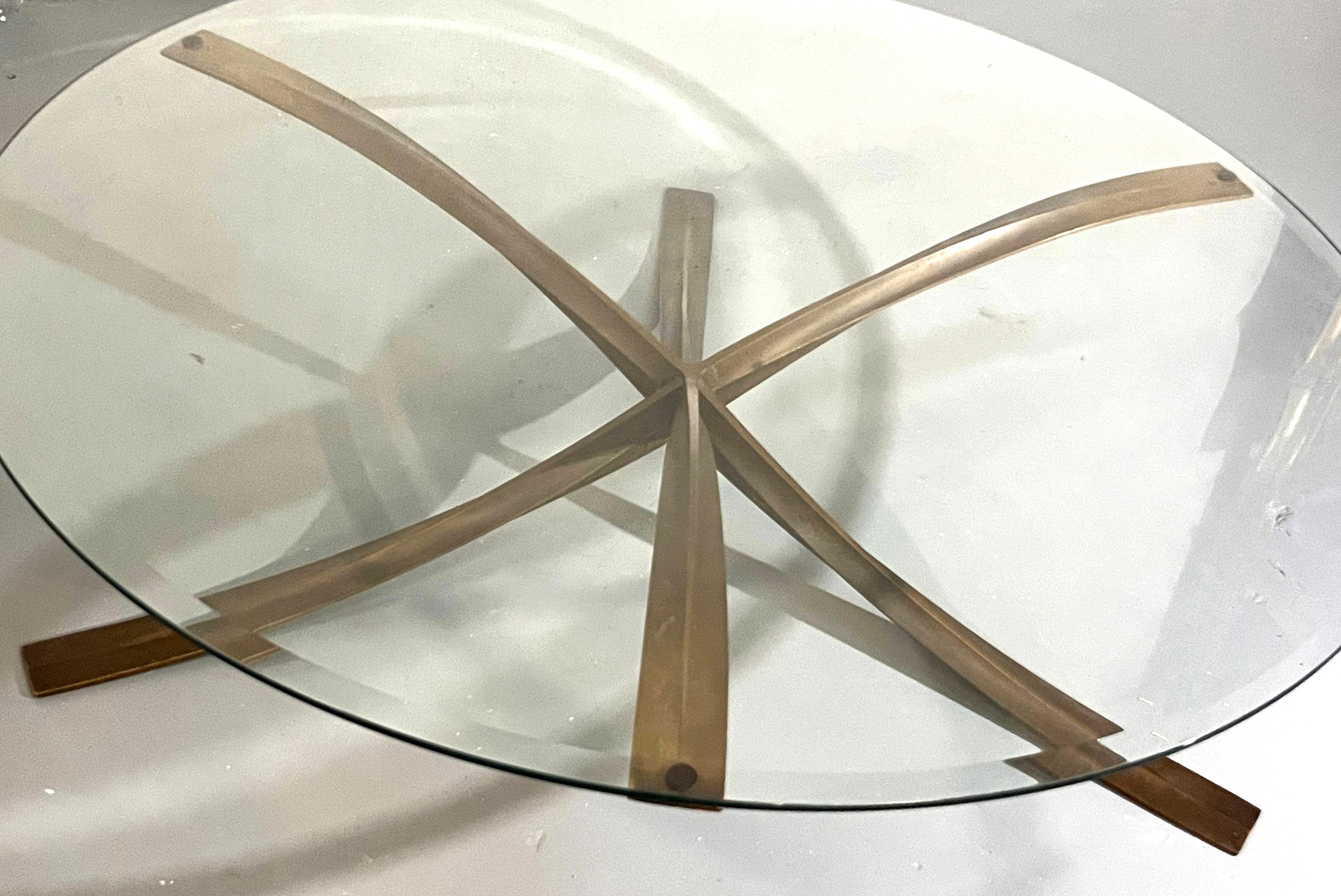 La mesa presenta una llamativa base escultórica compuesta por cuatro elementos curvos de bronce que se entrecruzan, formando una dinámica estructura estrellada. La cálida pátina del bronce contrasta maravillosamente con la tapa de cristal circular