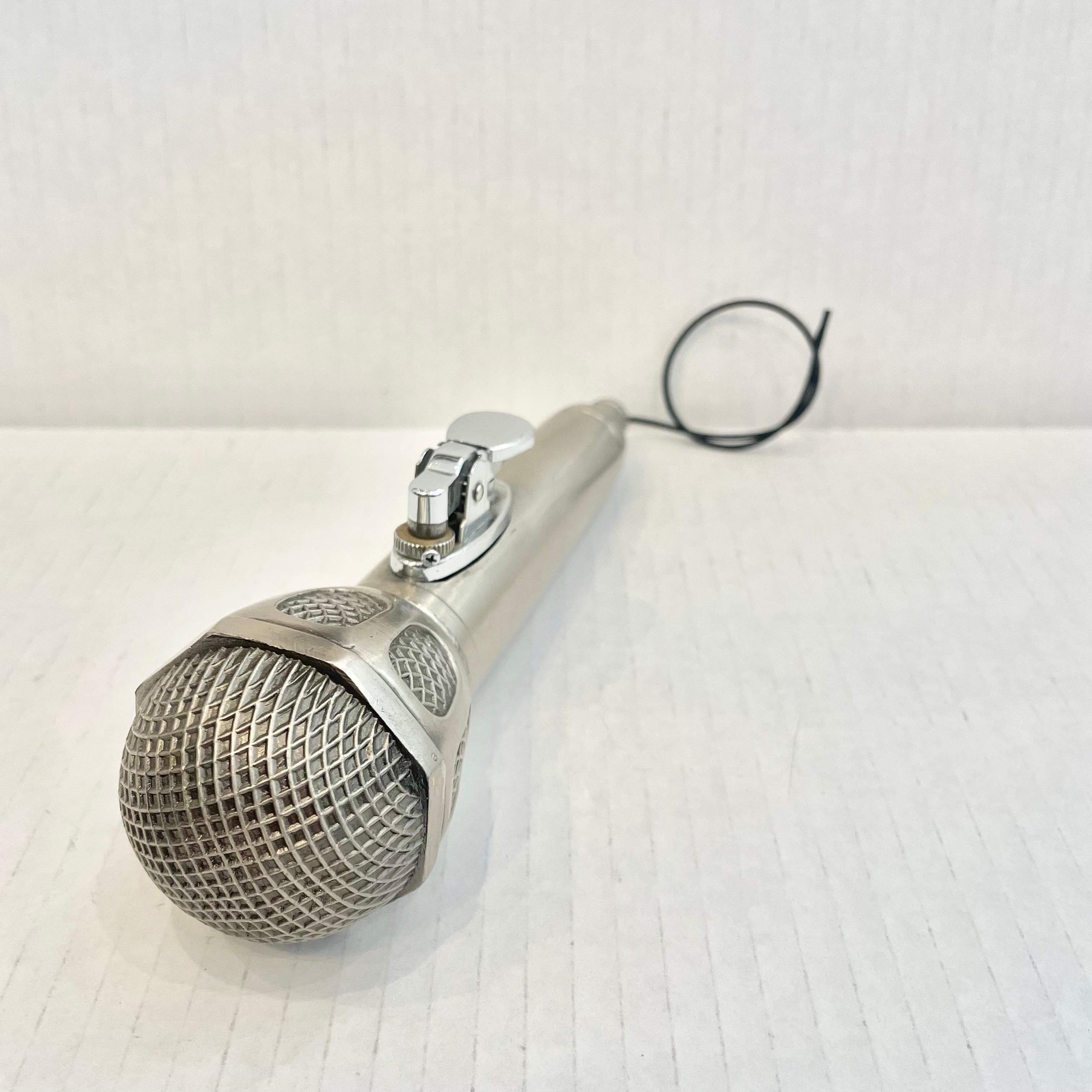 Great vintage table lighter in the shape of a microphone. Just over 7 inches long. Made completely of metal with a hollow body. Light patina giving this piece a classic silver color. The head has a detailed grille and metal casing. Small rubber cord