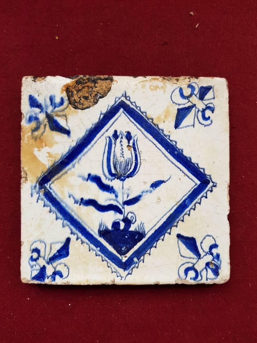 Mid-17th century Delft blue tile with a hand-painted decor of a flower in square and French lilies in every corner, it has a baking mistake upper left to the enamel what's inside.

The measurements are,
Depth 1.3 cm/ 0.5 inch.
Width 13 cm/ 5.1