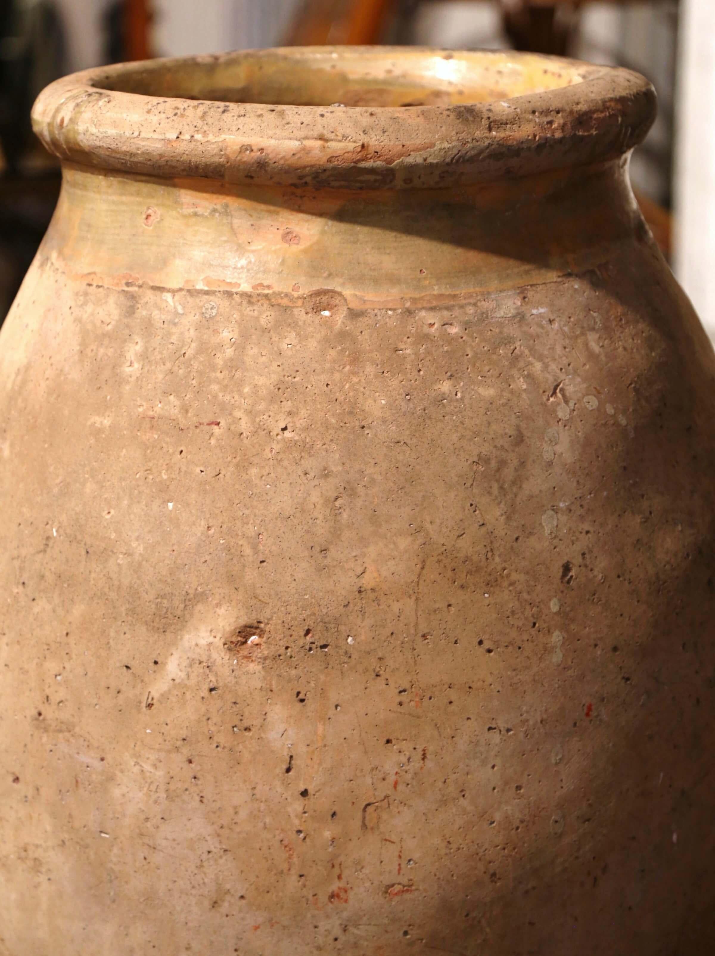 This large, antique earthenware olive jar was created in Southern France, circa 1760. Made of blond clay and neutral in color, the terracotta vase has a traditional round shape. The rustic, time-worn pot features a yellow glaze around the neck and