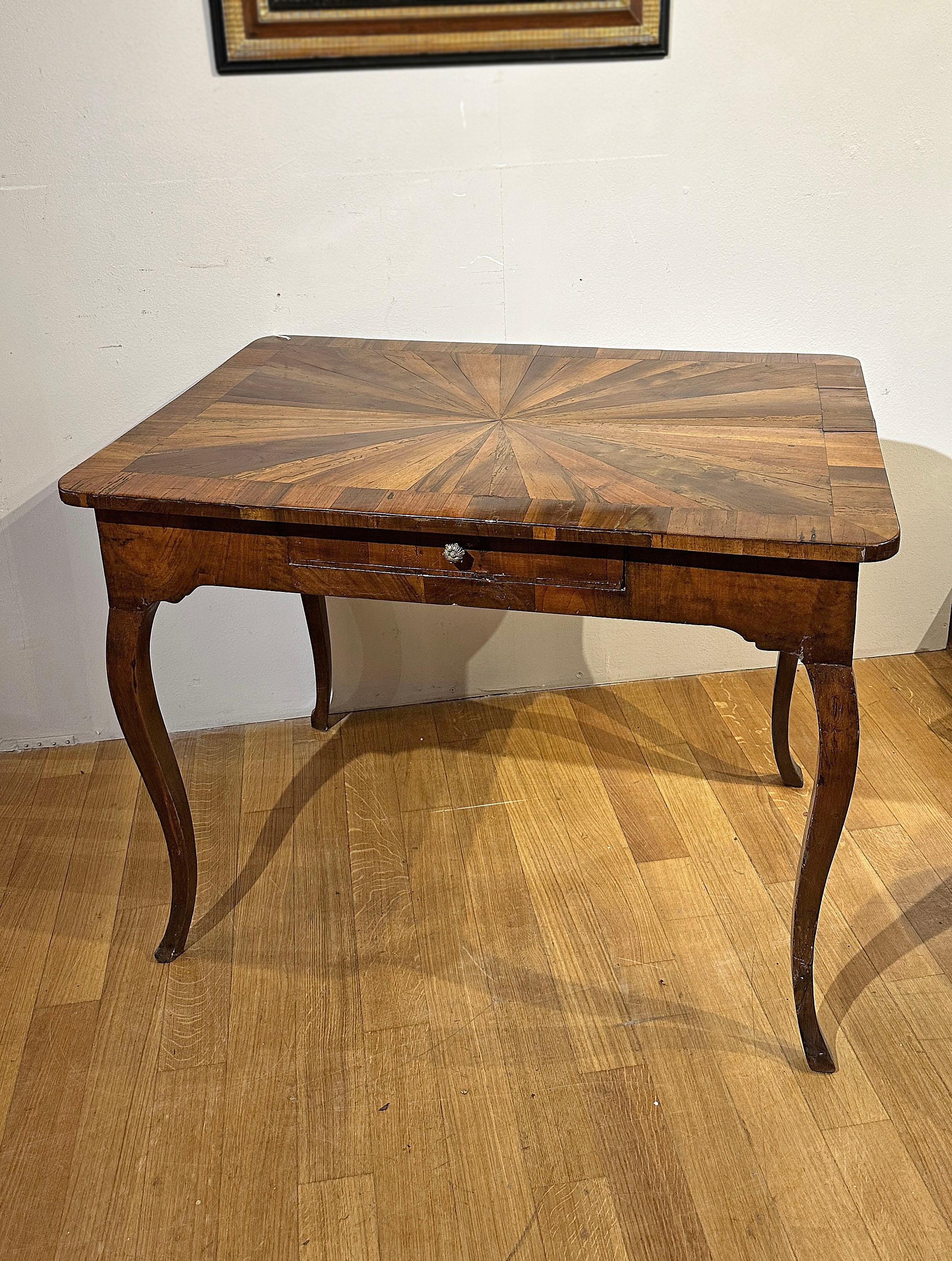 Élégante table à écrire entièrement plaquée de noyer et d'olivier, agrémentée d'incrustations sur le plateau. La table se distingue par ses pieds légèrement incurvés, typiques du style de l'époque, et par un tiroir central ergonomique, idéal pour