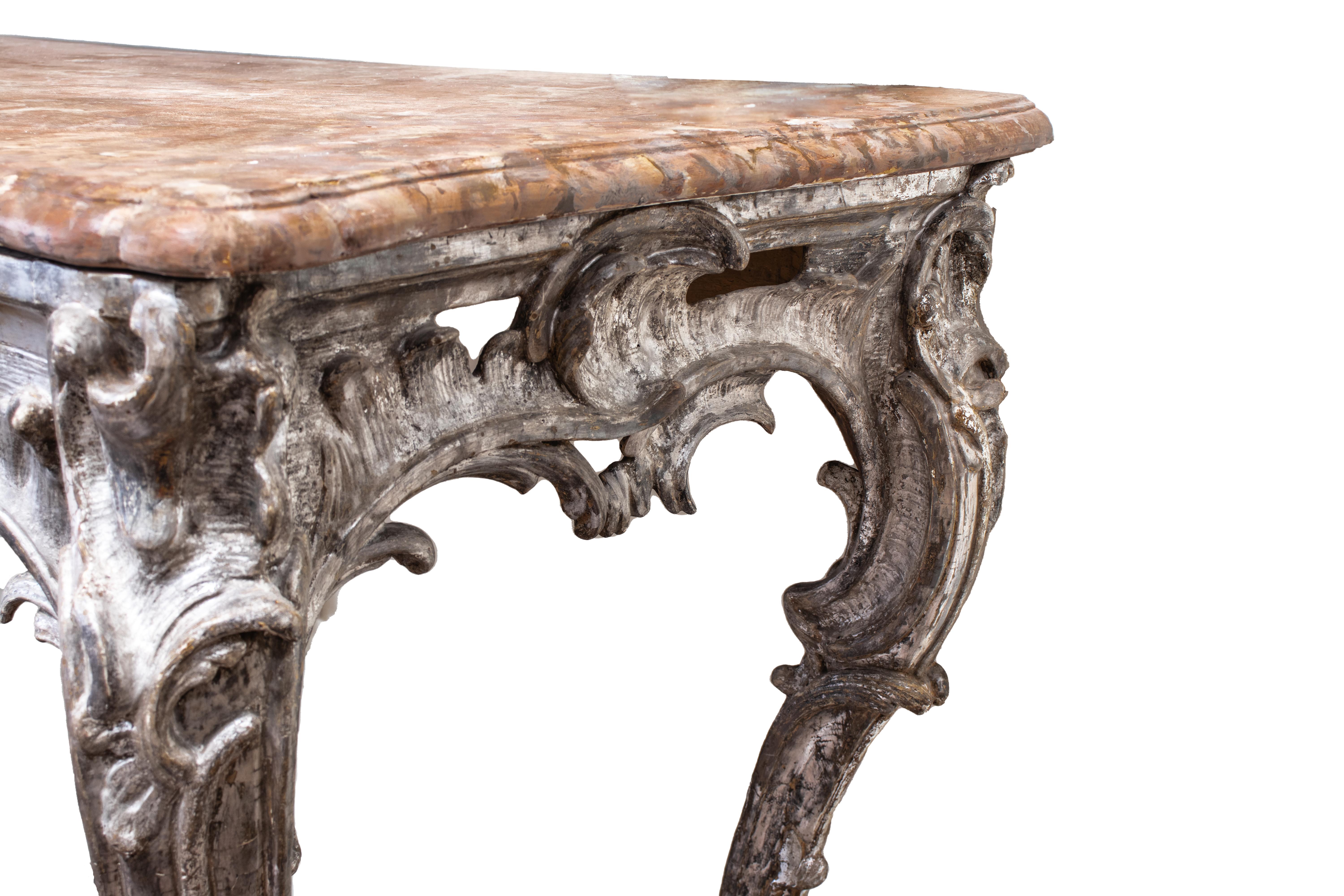 South German mid-18th-century carved wood console in Rococo style. The curved feet are adorned with rocaille scrolls and foliage motifs, and the frame is decorated with a massive floral element at the center. The console base is entirely silvered,